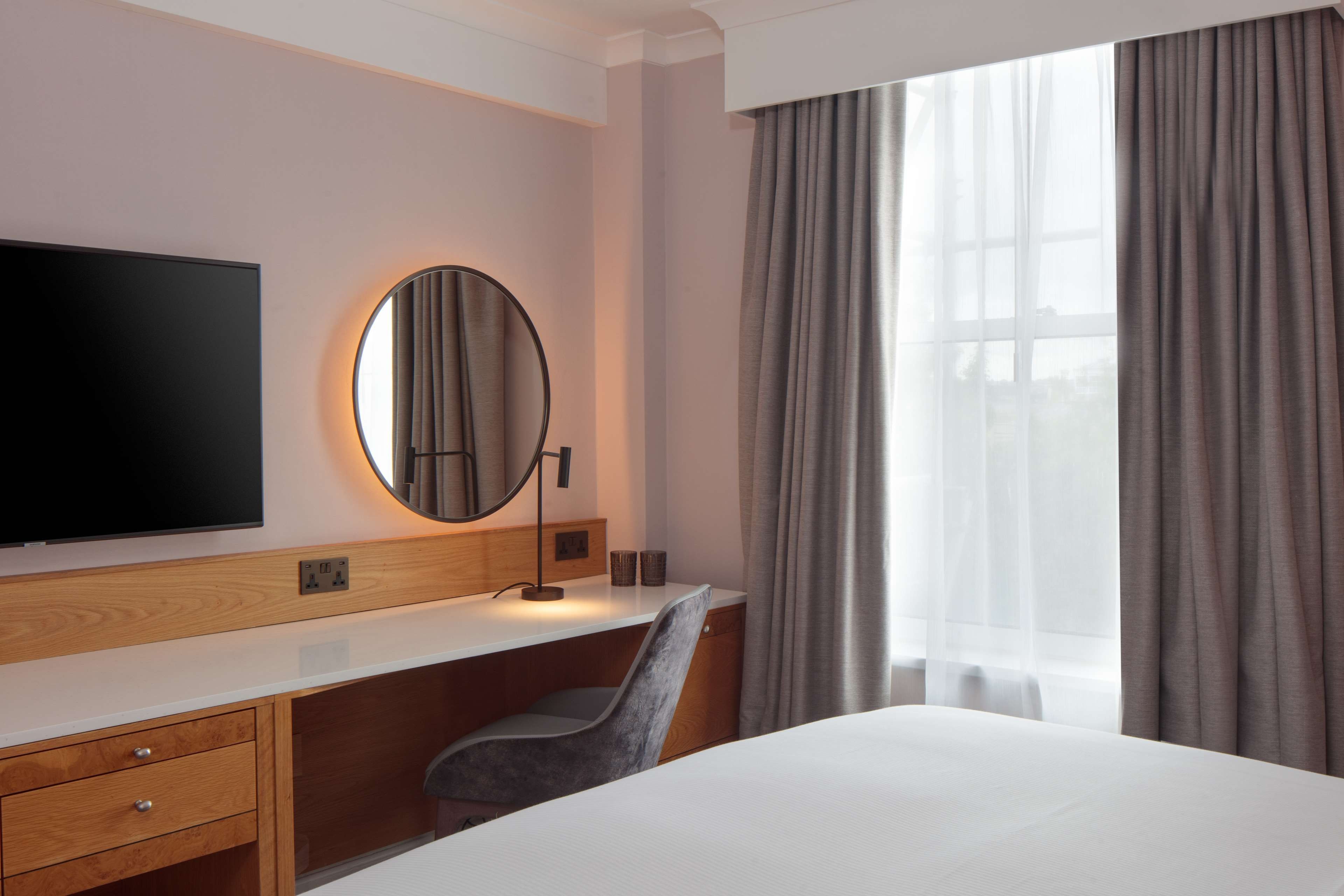 A hotel room with a large bed, a desk with a chair, a flat-screen TV, a round mirror, and a window with curtains.