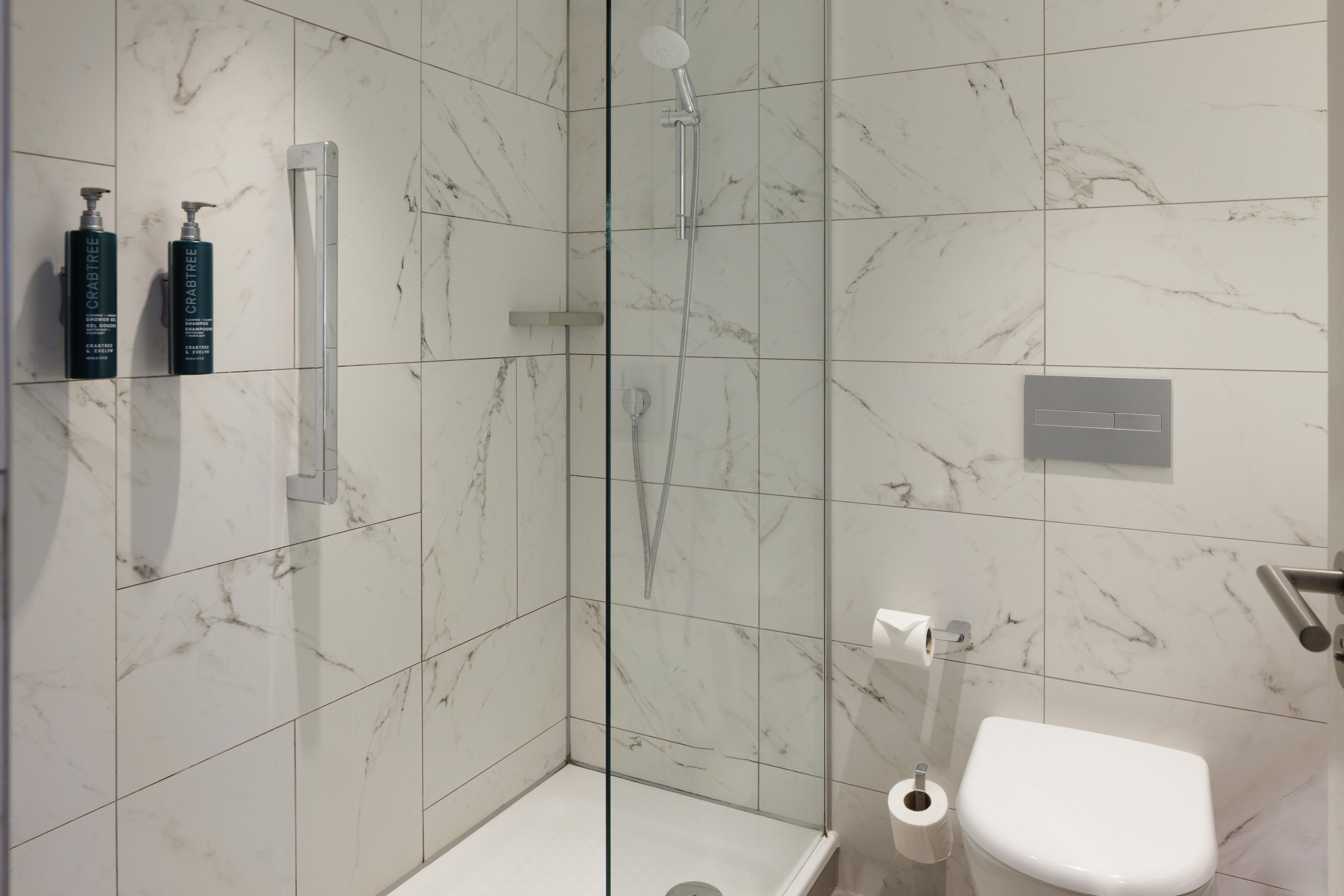 A modern bathroom with a glass-enclosed shower, marble tiles, a toilet, and two bottles of Crabtree & Evelyn shampoo and body wash.