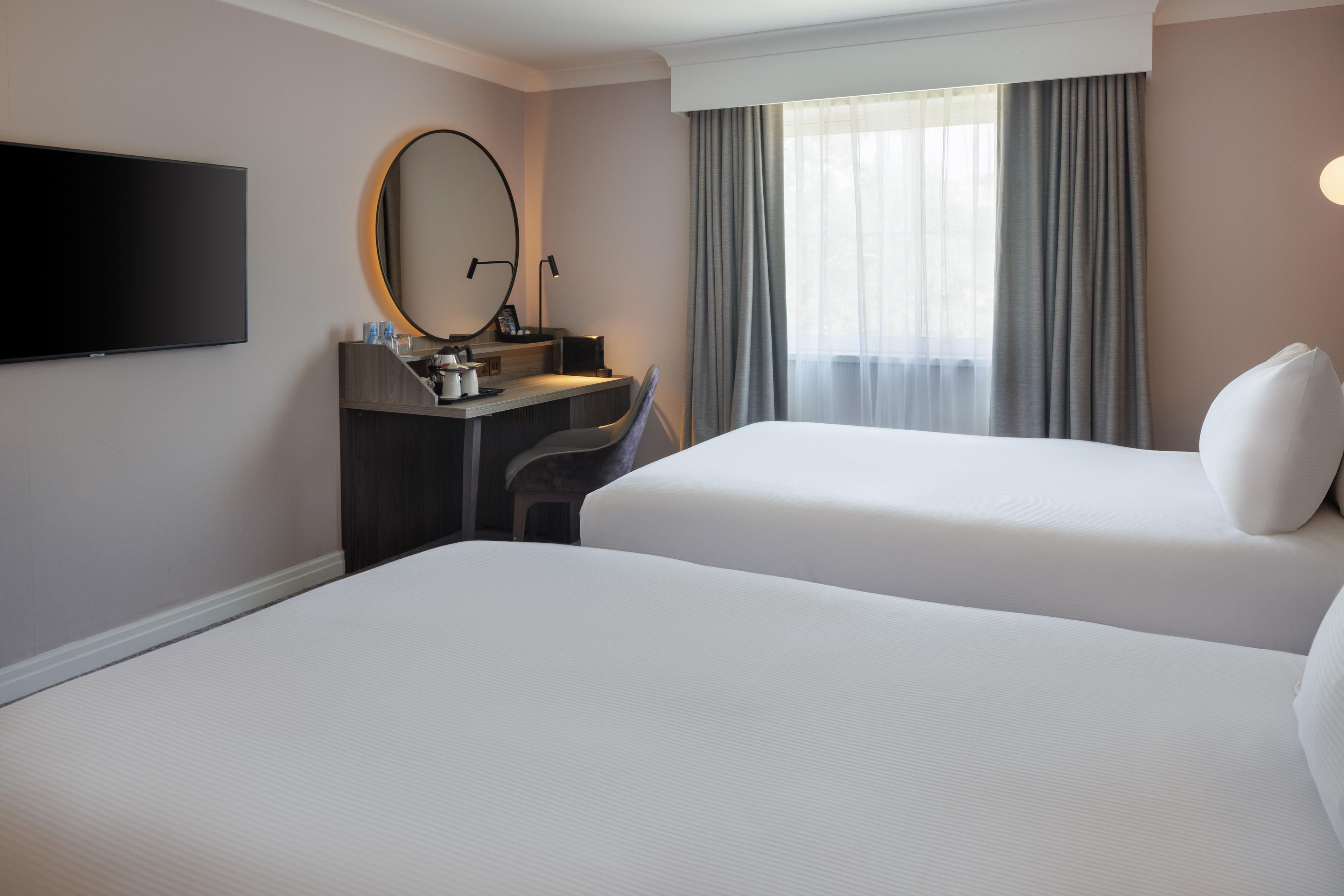 A hotel room with two beds, a desk with a chair, a flat-screen TV, and a large window with curtains.