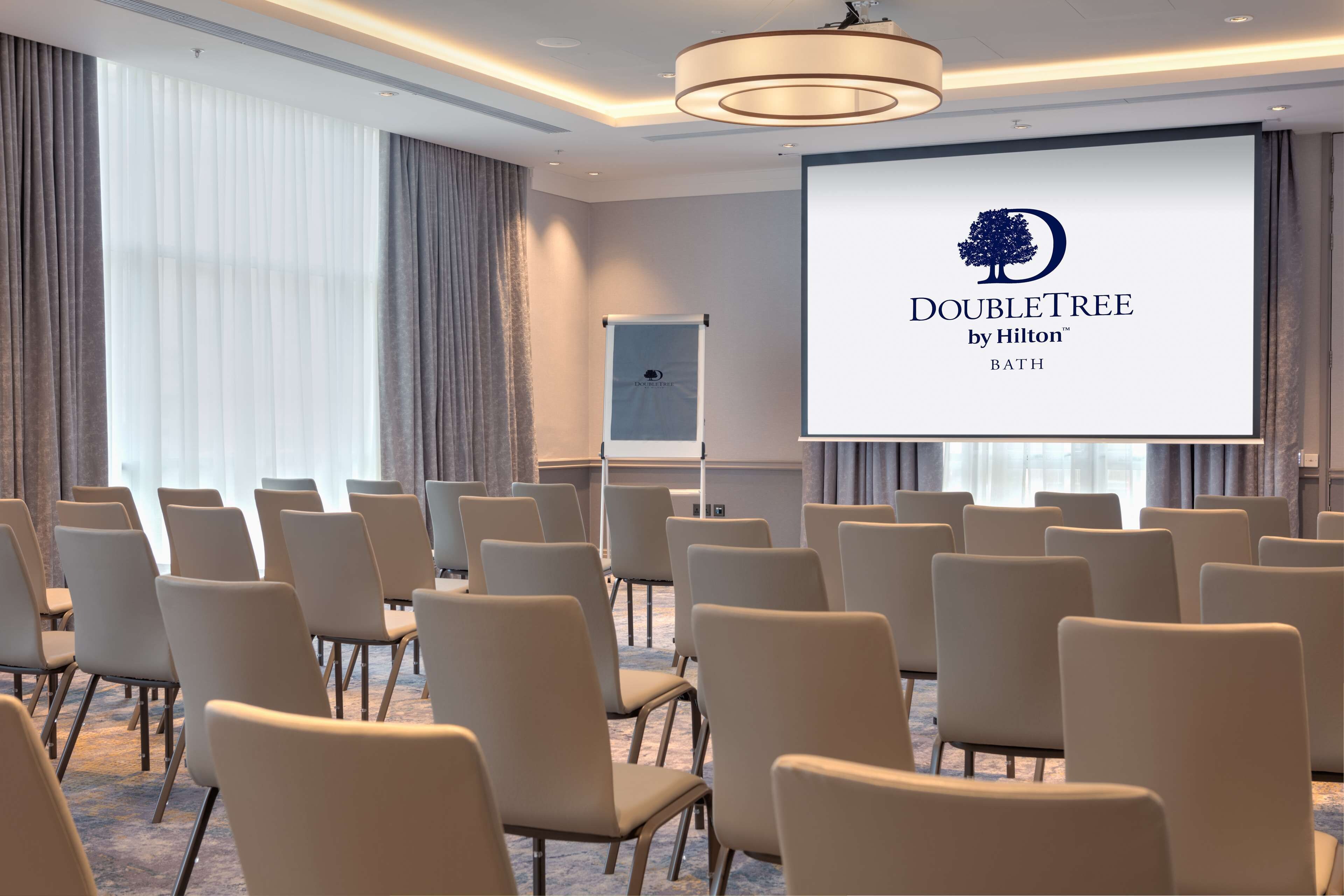 Uma sala de conferências com fileiras de cadeiras, uma tela de projetor exibindo o logotipo da DoubleTree by Hilton e um quadro branco.