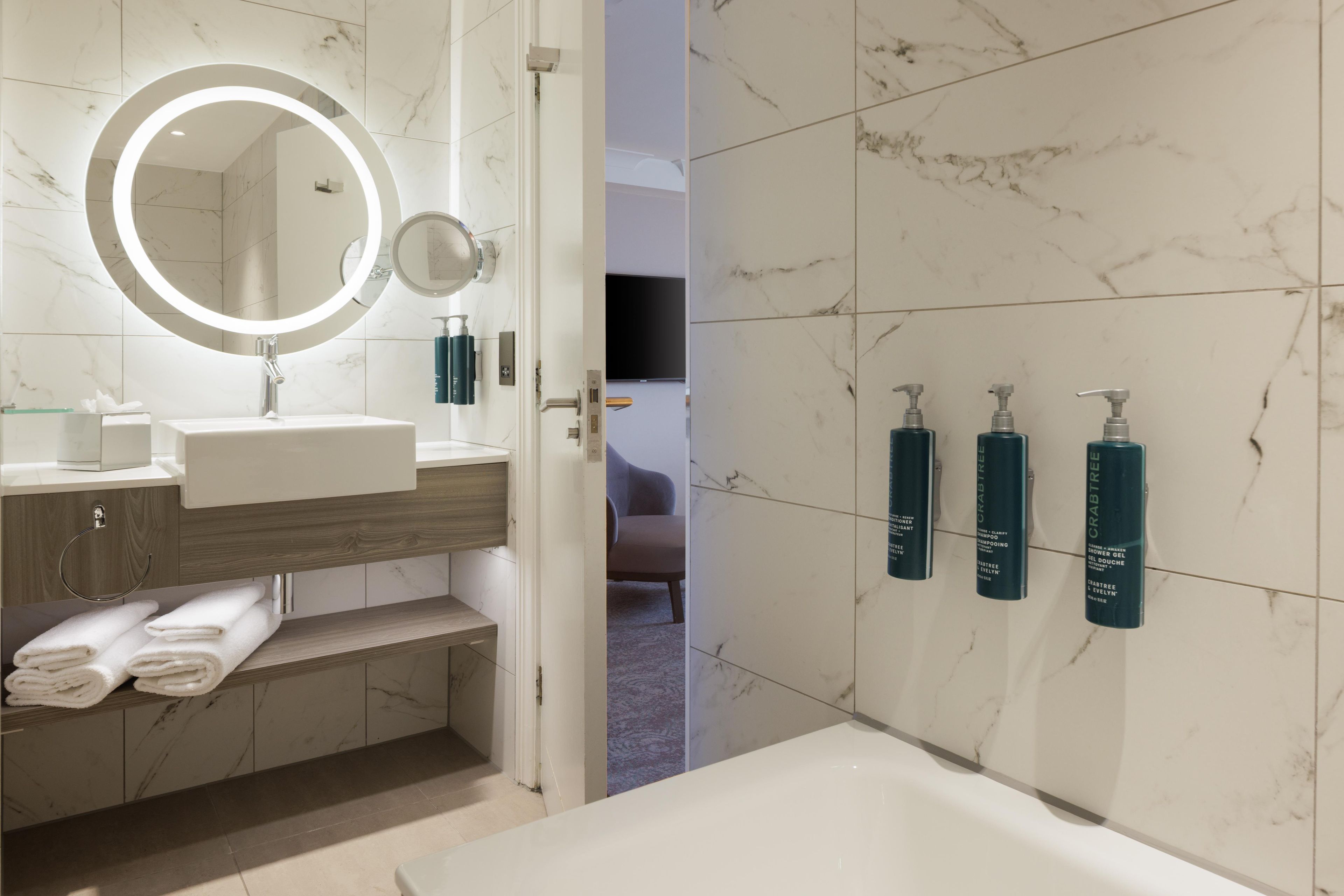 A modern bathroom with a marble-tiled wall, a round mirror, a sink, a vanity, and three bottles of Crabtree & Evelyn products.