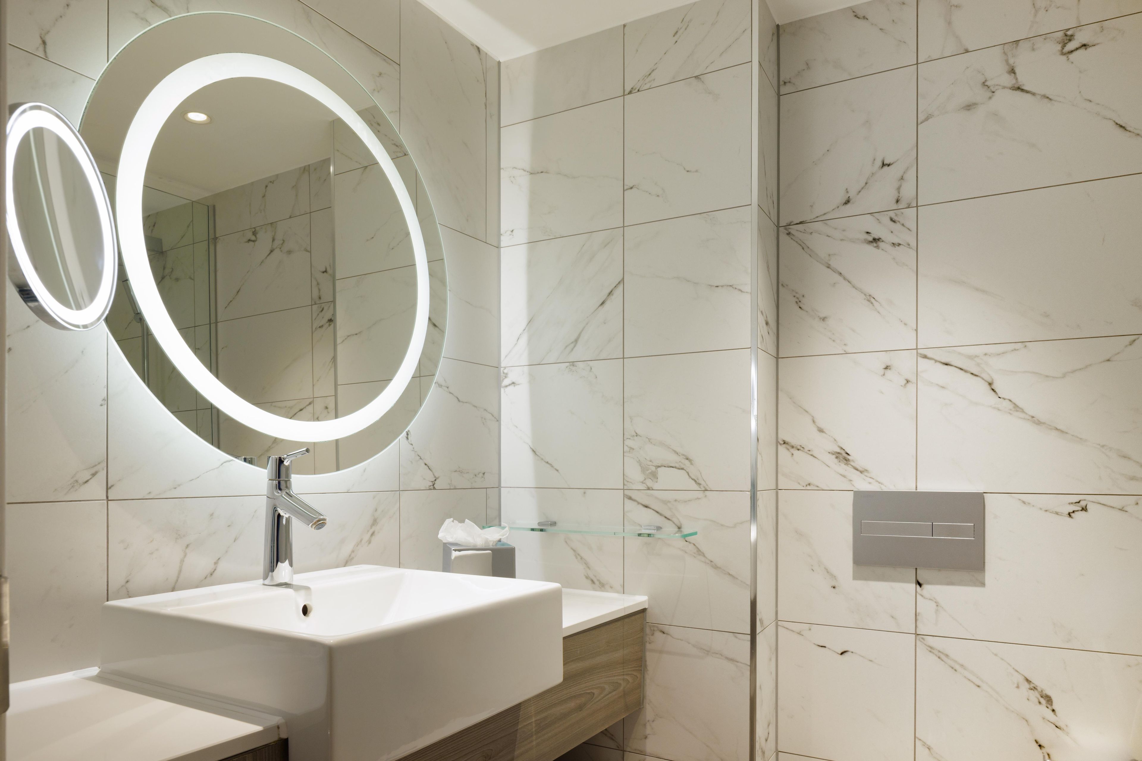 A modern bathroom with a double sink vanity, a large mirror with integrated lighting, and marble-patterned walls.