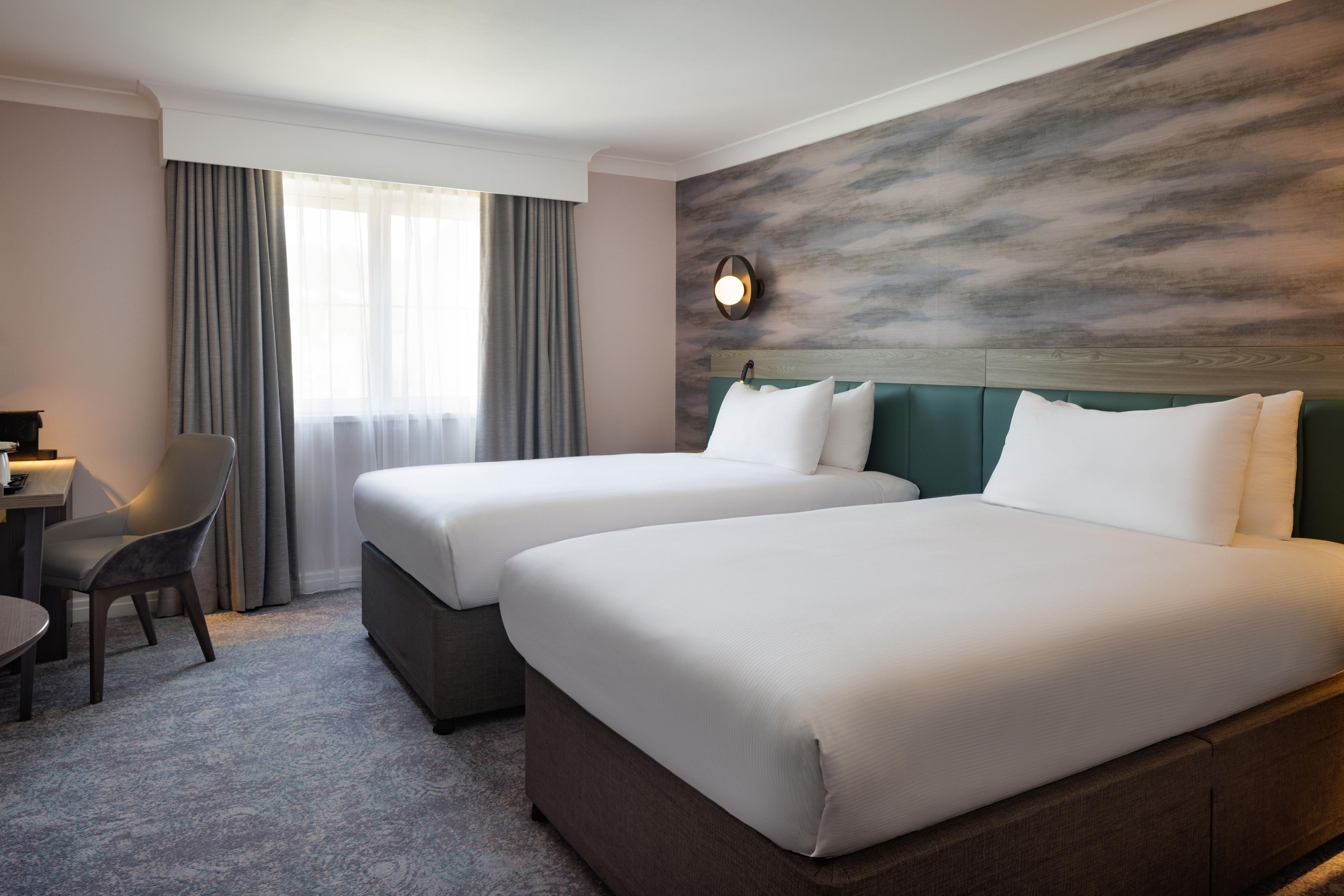 Una habitación de hotel con dos camas, un escritorio, una silla y una ventana con cortinas.