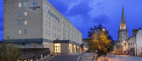 A hotel with a modern facade, a prominent entrance, and a church spire in the background.