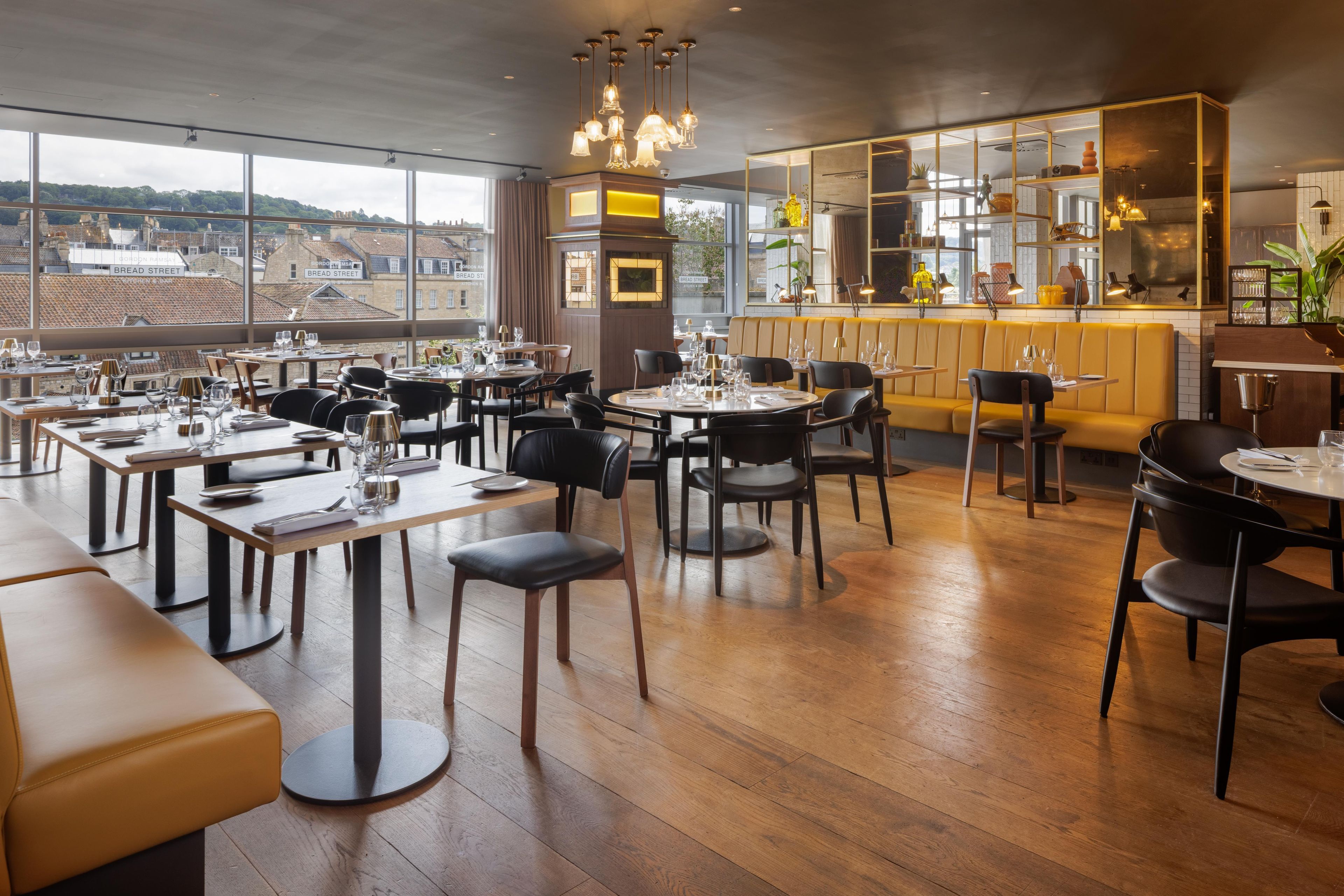A restaurant with multiple dining tables and chairs, large windows offering a view of the city, and a bar area with shelves and plants.