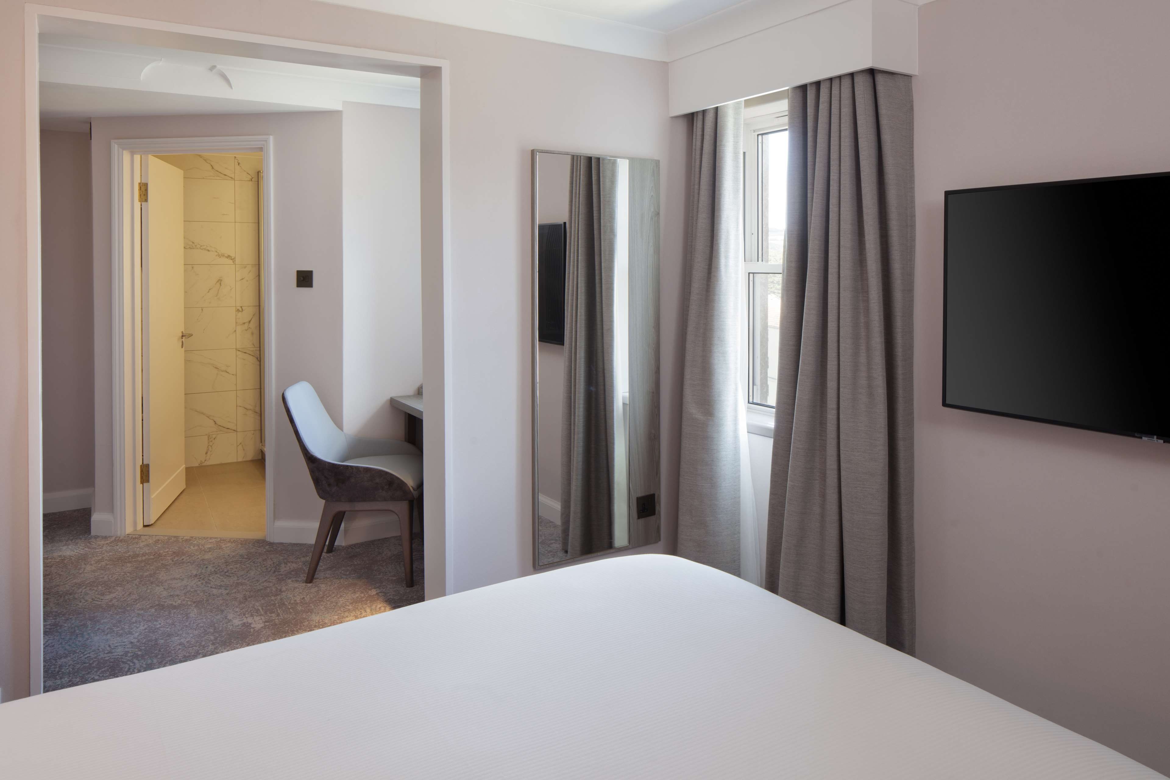 Habitación de hotel con cama, escritorio, silla, televisión y baño visible a través de una puerta abierta.