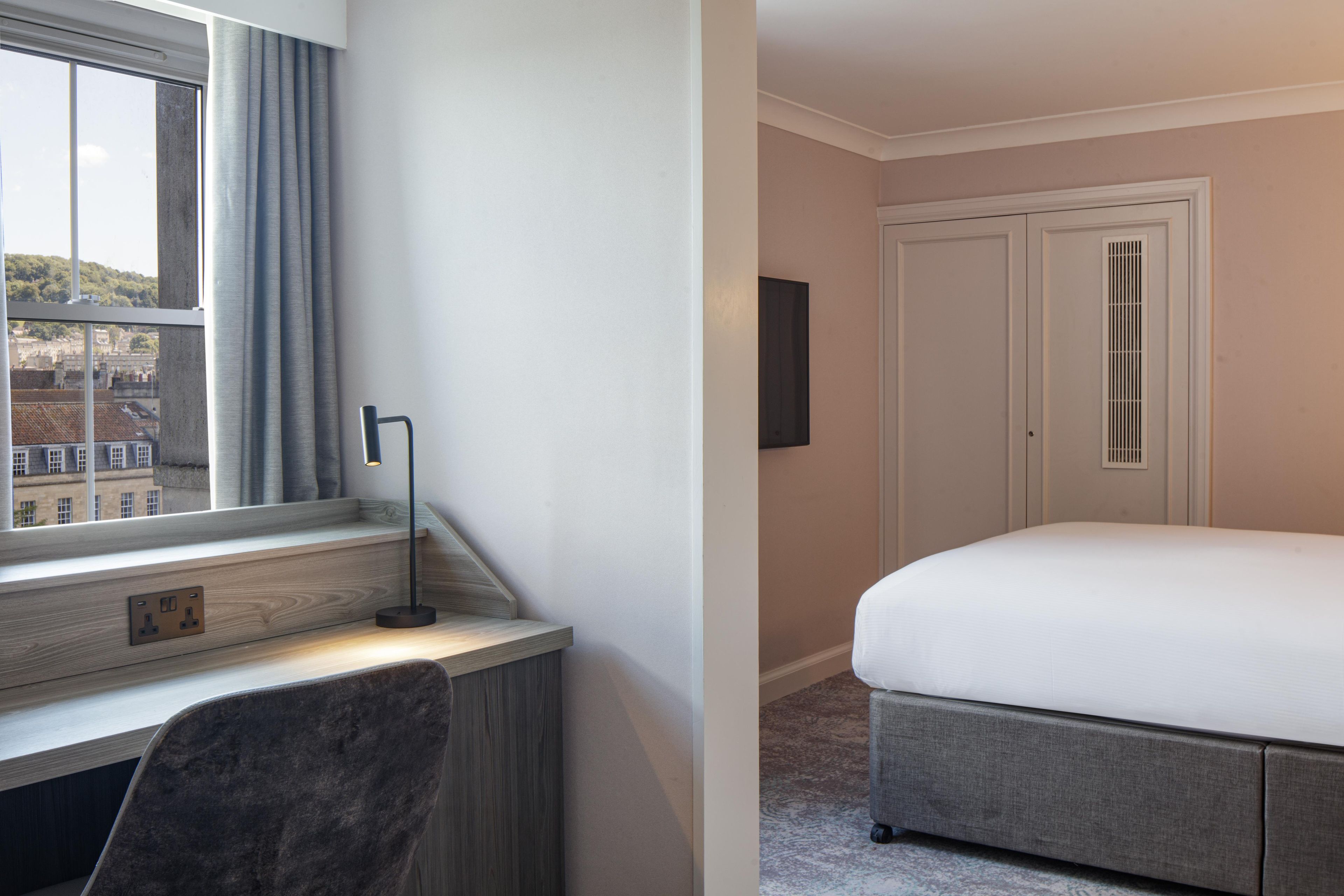 A hotel room with a bed, a desk with a chair, a window with curtains, and a door.