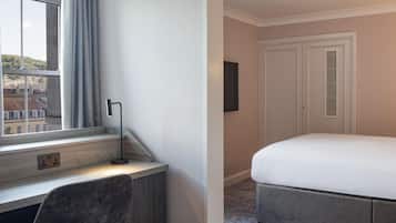 A hotel room with a bed, a desk with a chair, a window with curtains, and a door.