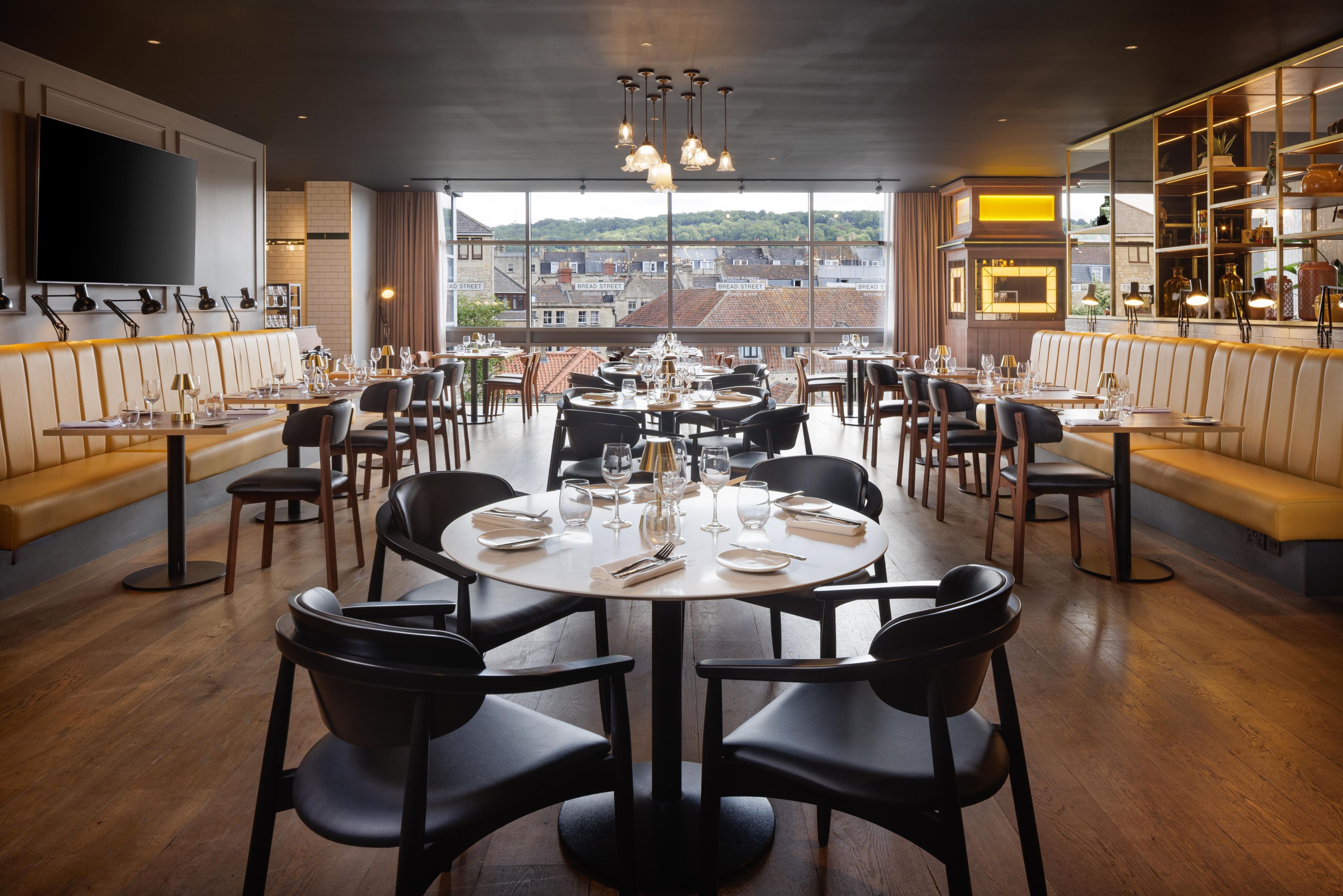 Un restaurante con mesas redondas, sillas negras y vistas a edificios a través de amplios ventanales.