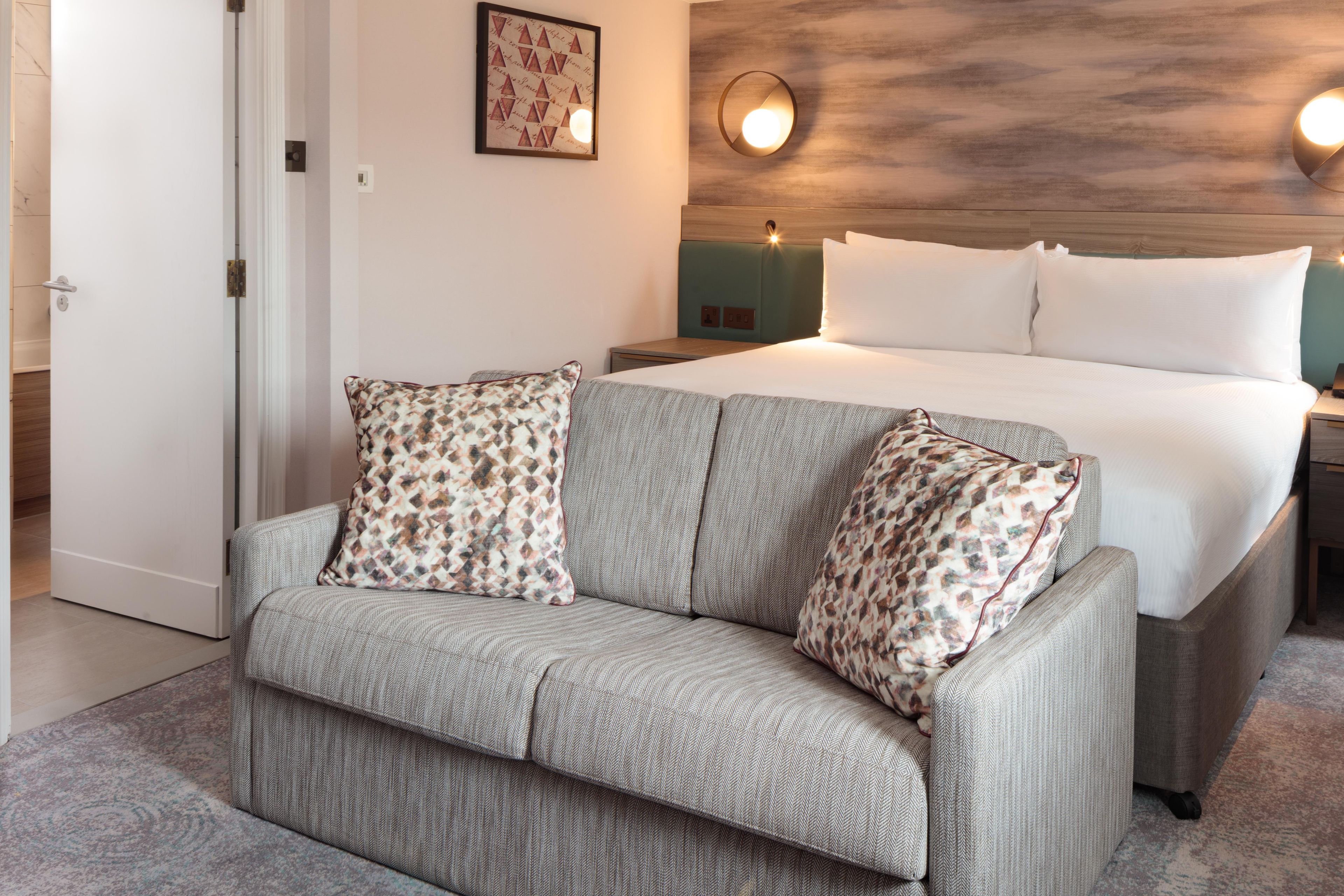 A hotel room with a bed, a sofa, and a wall-mounted light fixture.