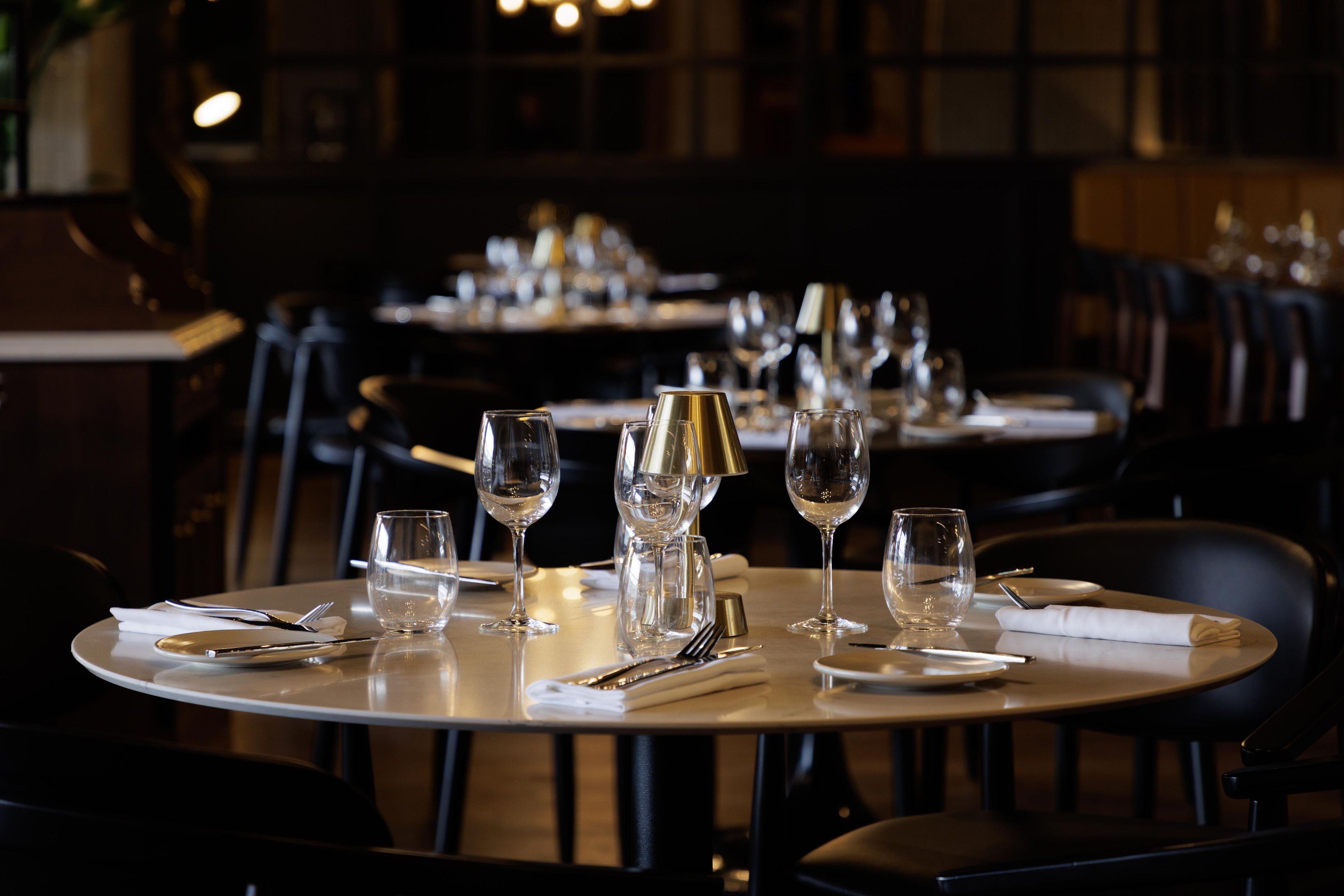 A restaurant setting with round tables, each set with plates, cutlery, and wine glasses.