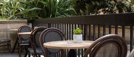 An outdoor seating area with wooden tables and chairs, surrounded by greenery and a stone wall.