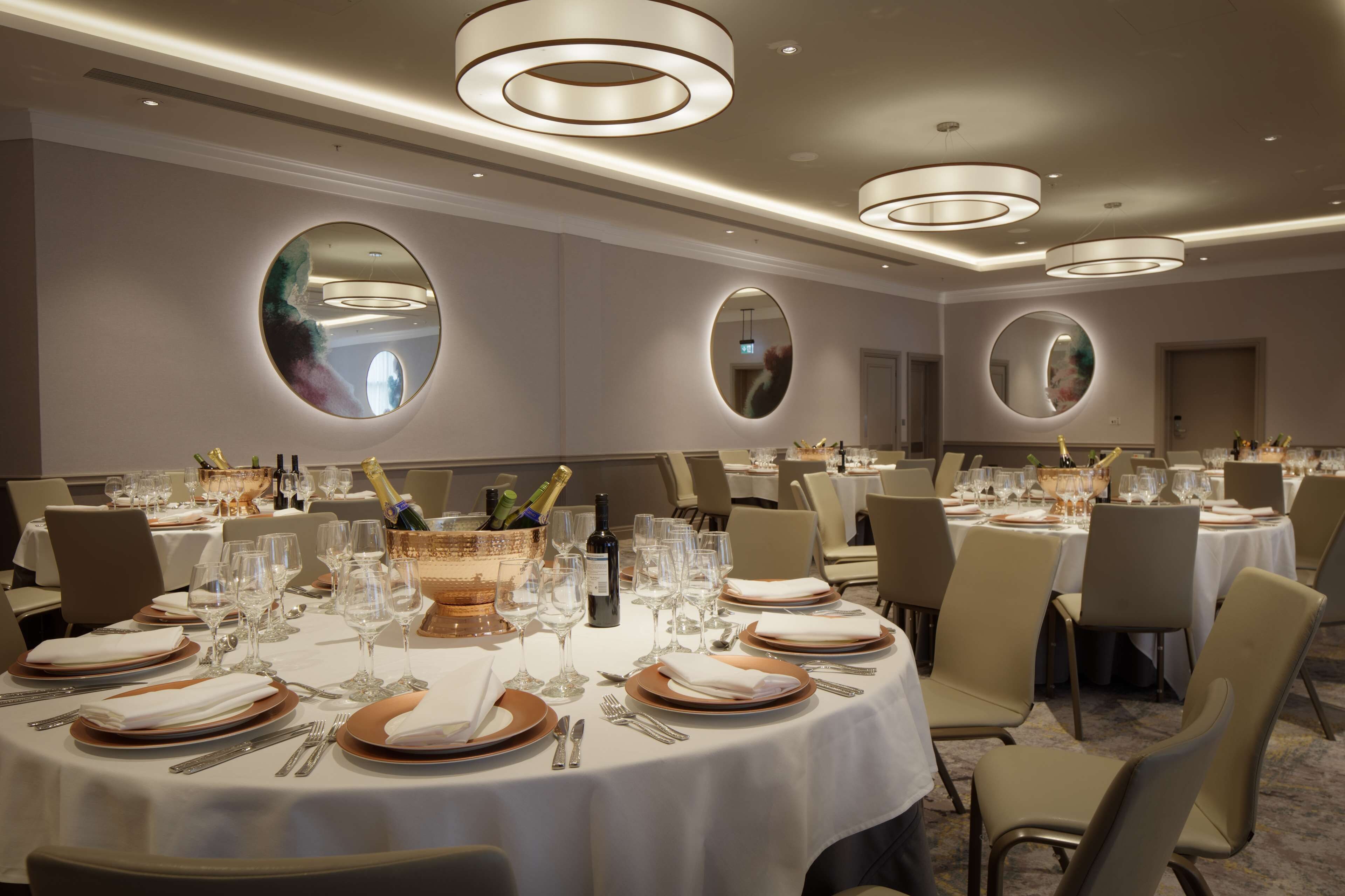 Un salón de banquetes con mesas redondas dispuestas para cenar, con manteles blancos, platos con detalles dorados y copas de vino.
