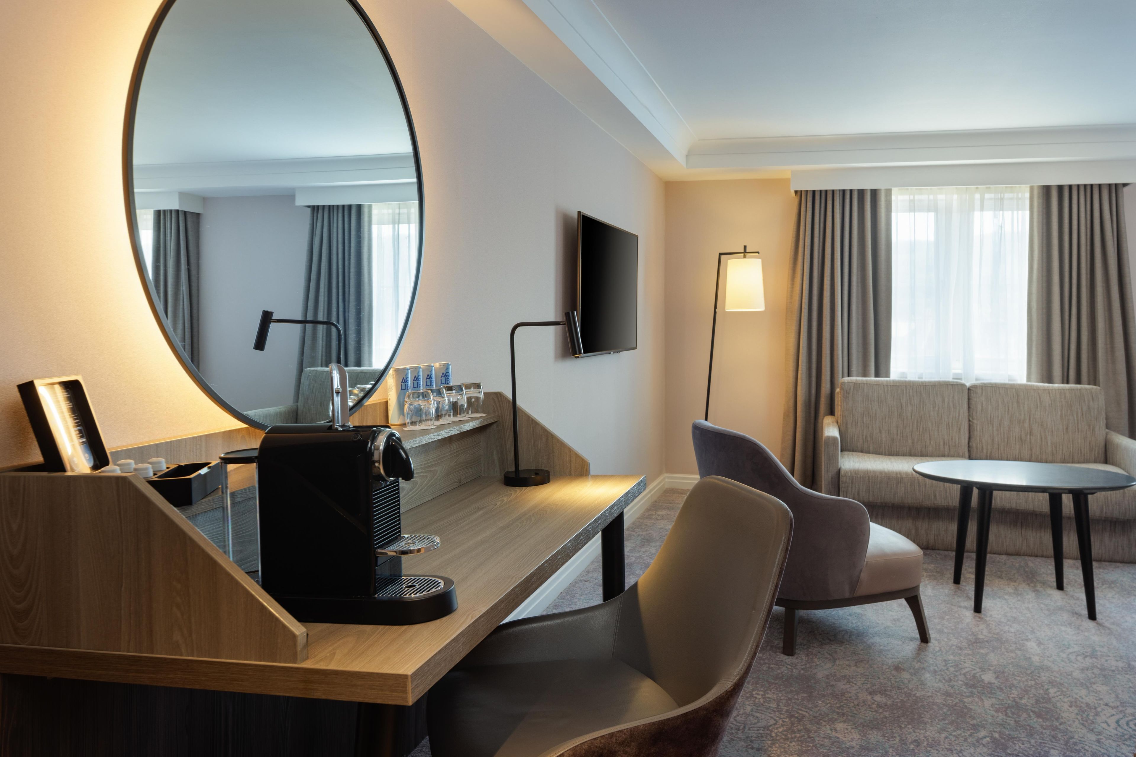 A hotel room with a wooden desk, a round mirror, a flat-screen TV, a sofa, a small table, and a coffee machine.