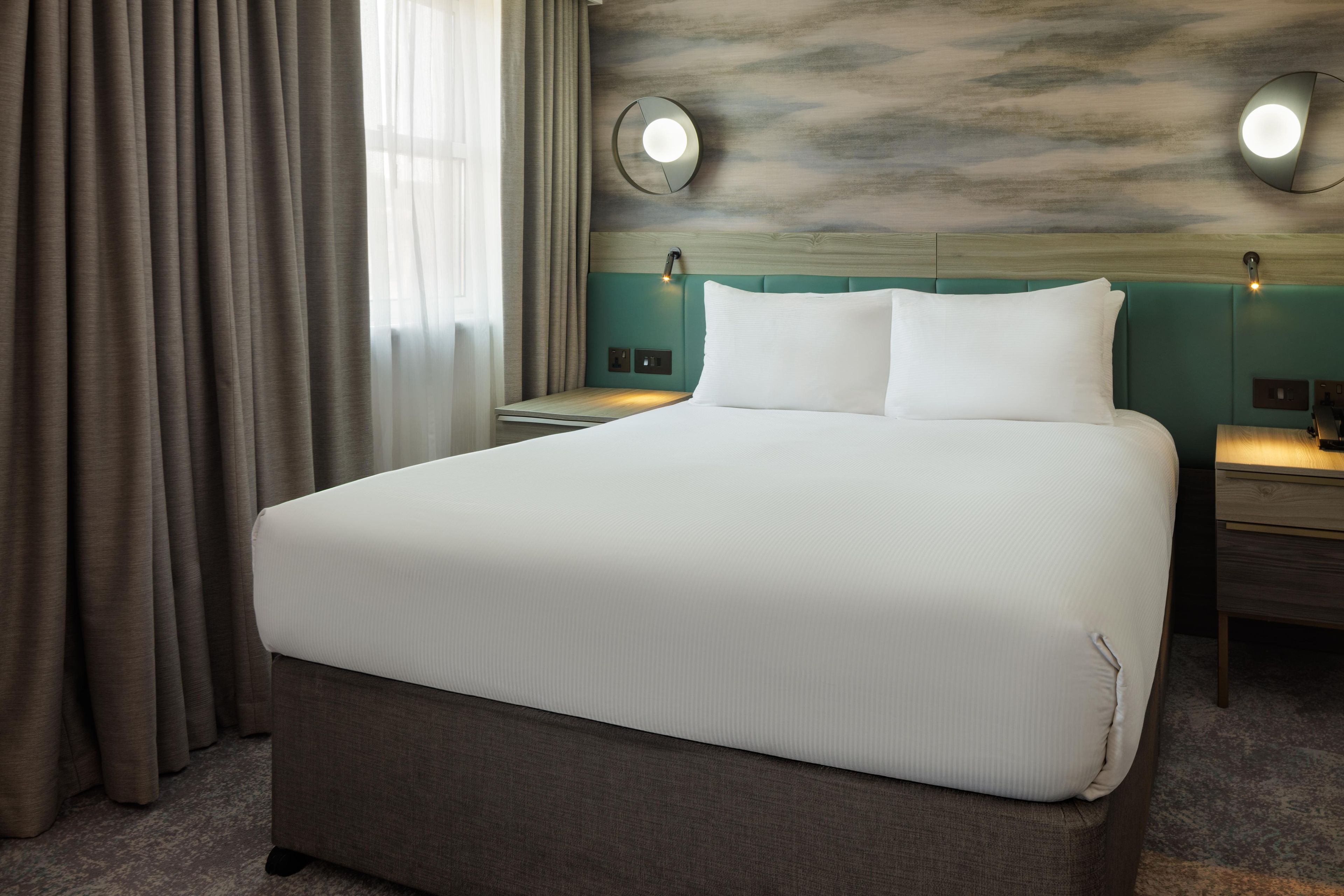A hotel room with a large bed, two bedside tables, a headboard with integrated lighting, and a view of the sky through the window.