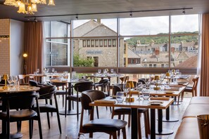 A restaurant with tables set for dining, large windows offering a view of the city, and a brick wall on one side. - Doubletree by Hilton Bath (Bath)