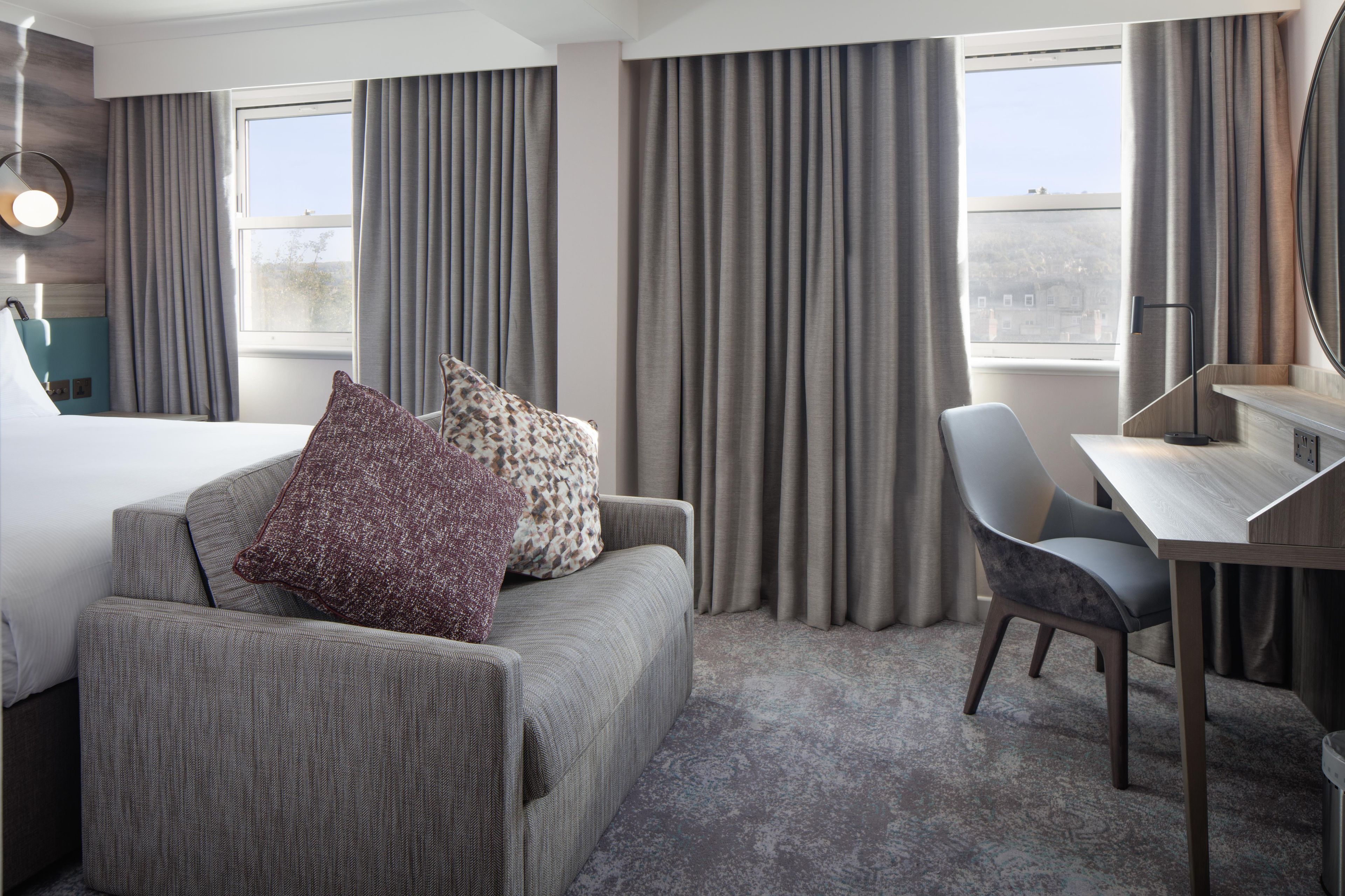 A hotel room with a bed, a sofa with two pillows, a desk with a chair, and large windows with curtains.