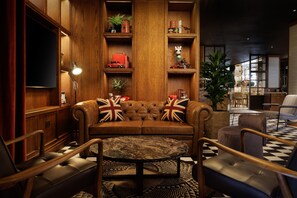 A cozy lounge area with a leather sofa, armchairs, a round coffee table, and wooden shelving with various items. - Doubletree by Hilton Bath (Bath)