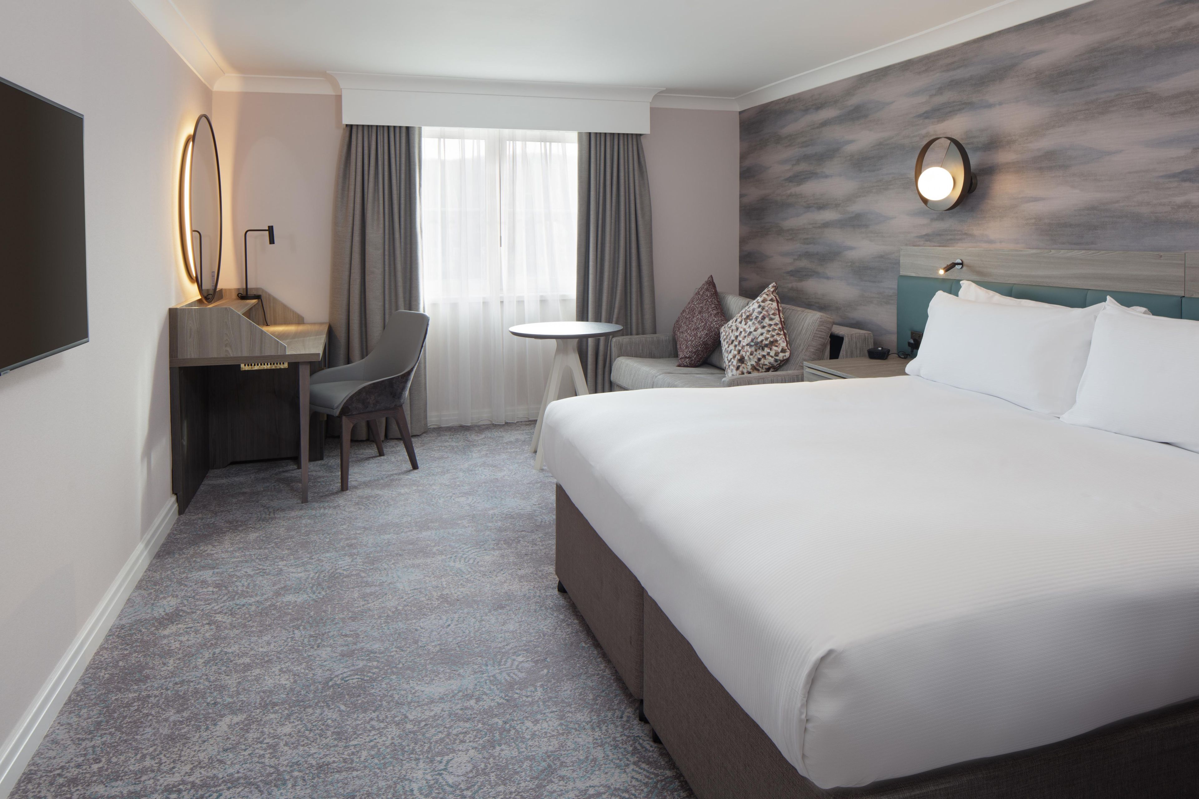 A hotel room with a large bed, a desk with a chair, a small round table, and a wall-mounted TV.
