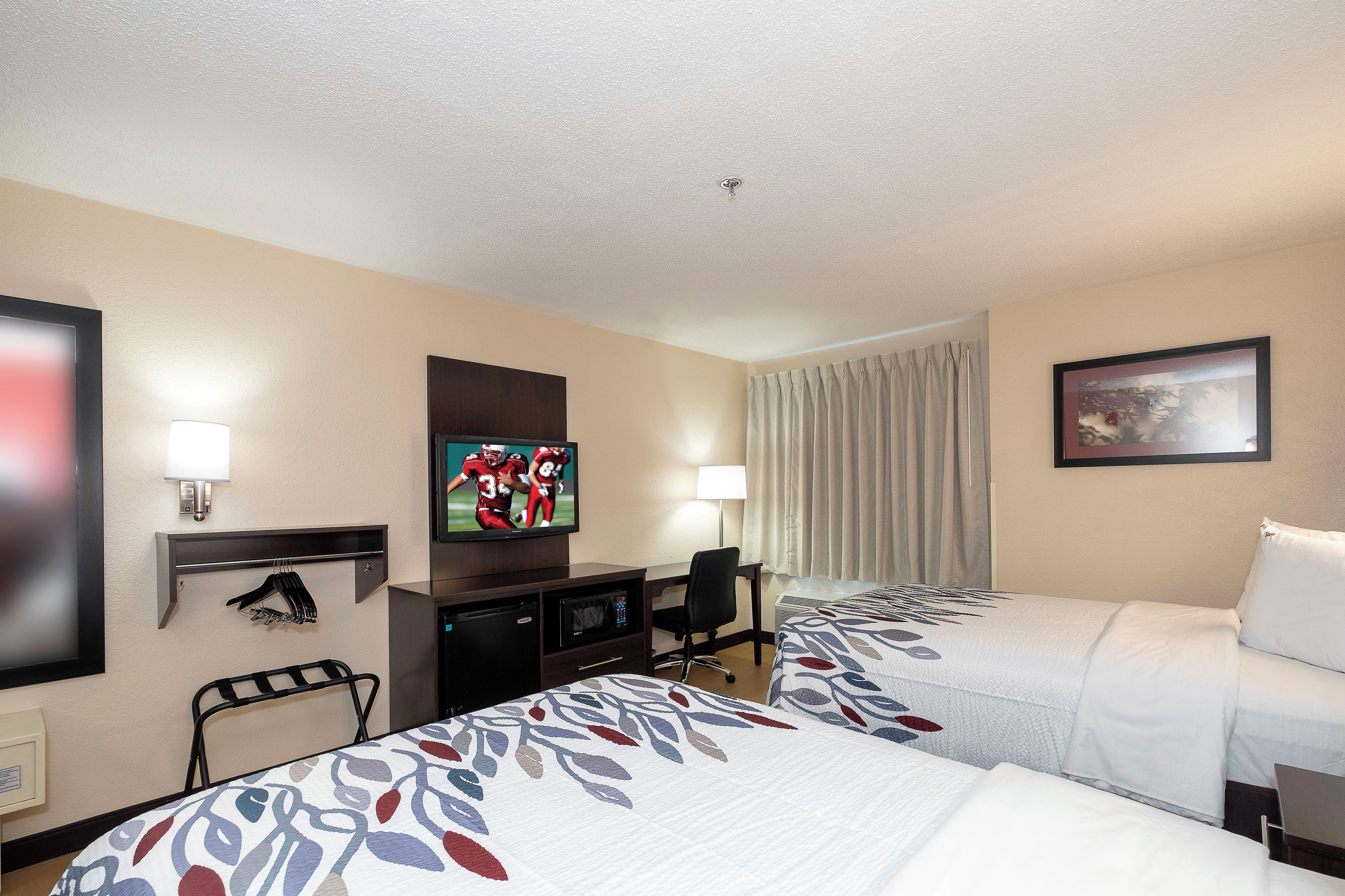 Ein Hotelzimmer mit zwei Betten, einem Fernseher, einem Schreibtisch mit Computer und einem Fenster mit Vorhängen.