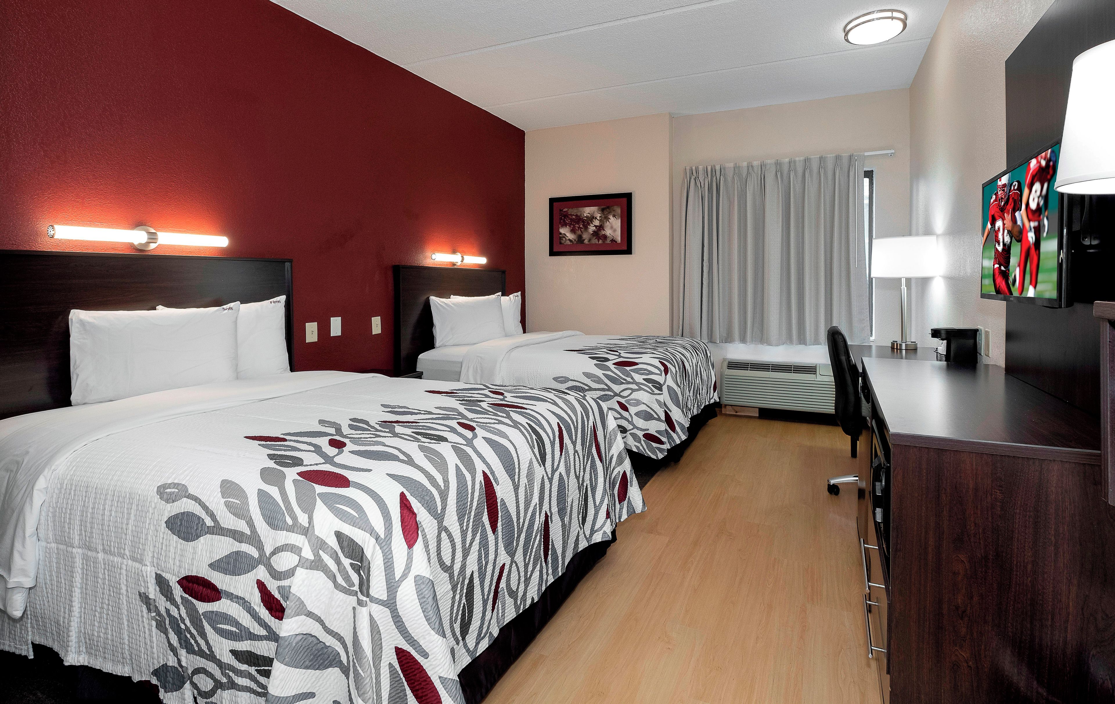 Quarto de hotel com três camas, uma escrivaninha com televisão e uma janela com cortinas.