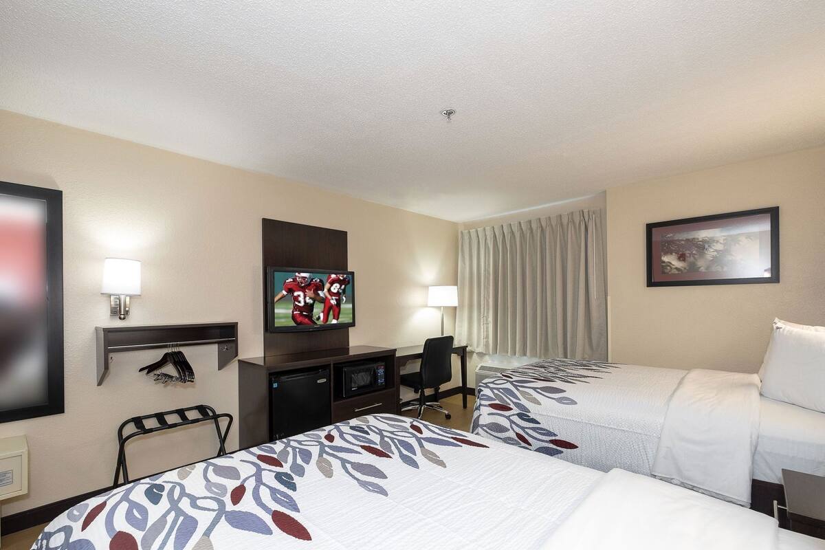 A hotel room with two beds, a desk, a television, a coat rack, and a window with curtains.
