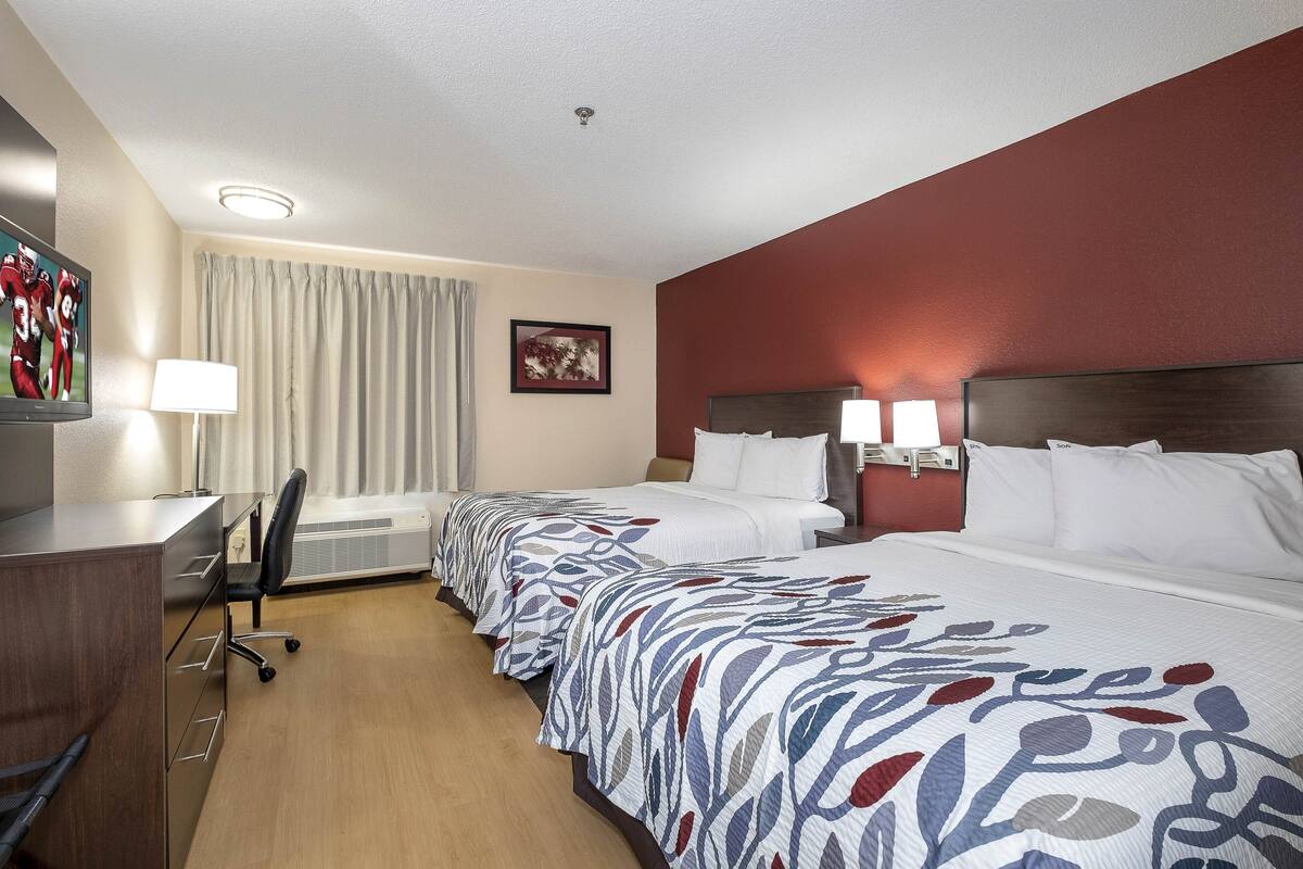 A hotel room with a desk, two beds, a chair, a lamp, a picture on the wall, and a window with curtains.