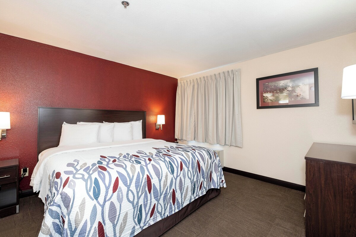 A hotel room with a large bed, a headboard, two bedside lamps, a nightstand, a desk, a chair, a window with curtains, and a framed picture on the wall.