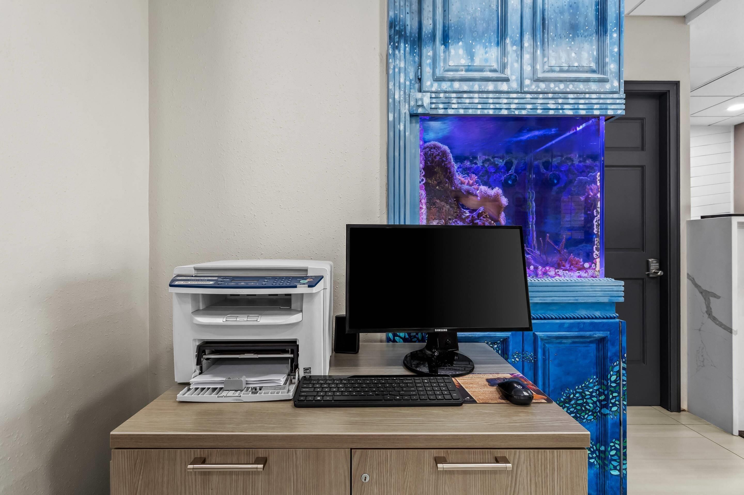 Un bureau avec un ordinateur, un clavier et une imprimante, un aquarium bleu en arrière-plan et une porte ornée d’un panneau bleu décoratif.
