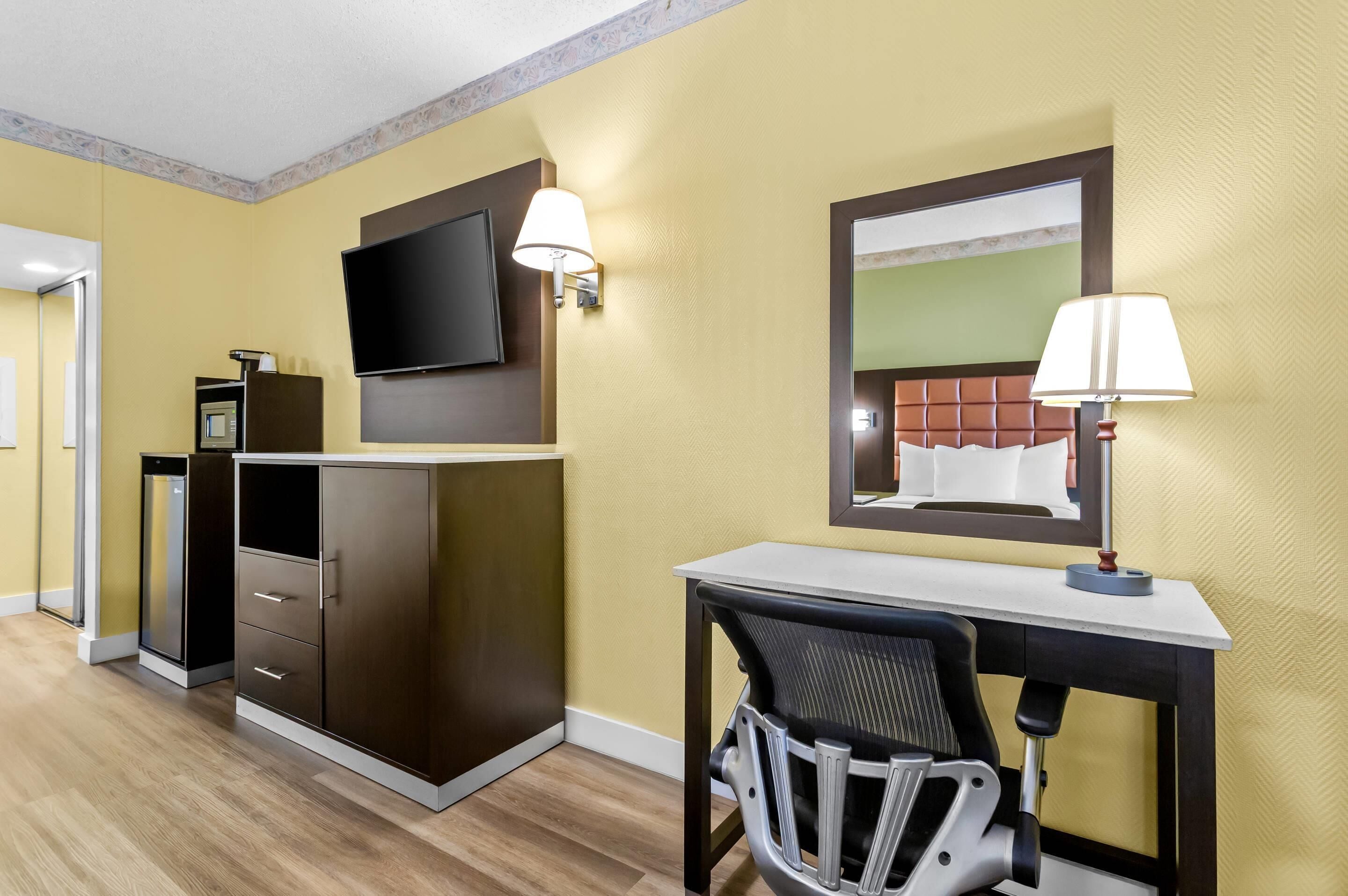 A hotel room with a desk, chair, television, wardrobe, and a mirror.