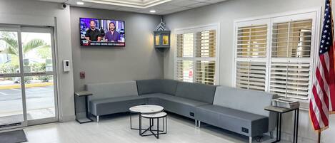 A reception area with a grey sectional sofa, a small round table, a TV mounted on the wall, and an American flag.