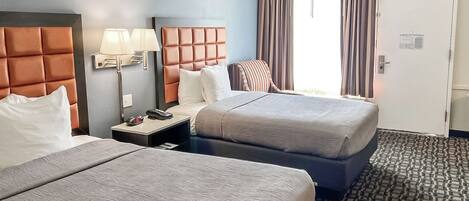 A hotel room with two beds, a nightstand with a lamp, a striped chair, and a door with a sign.