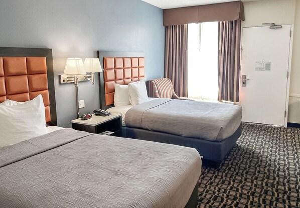 A hotel room with two beds, a nightstand with a lamp, a striped chair, and a door with a sign.