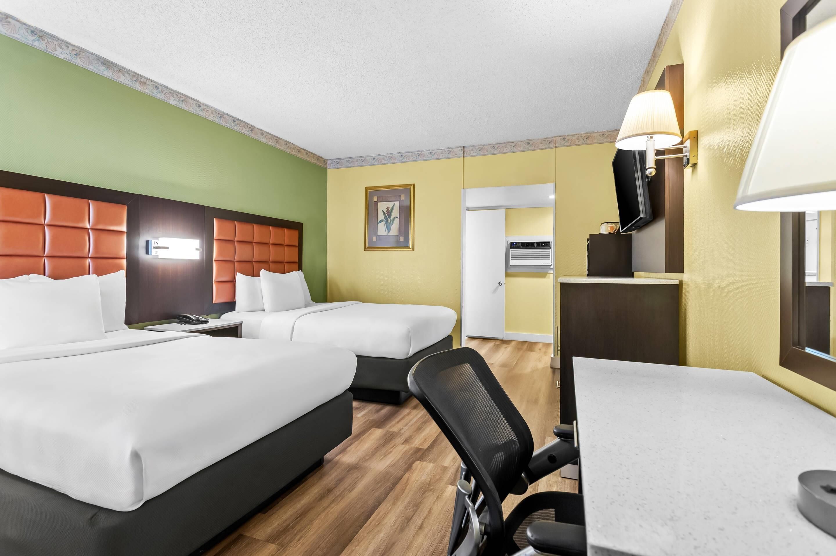 A hotel room with two beds, a desk, a chair, a TV, and a lamp.