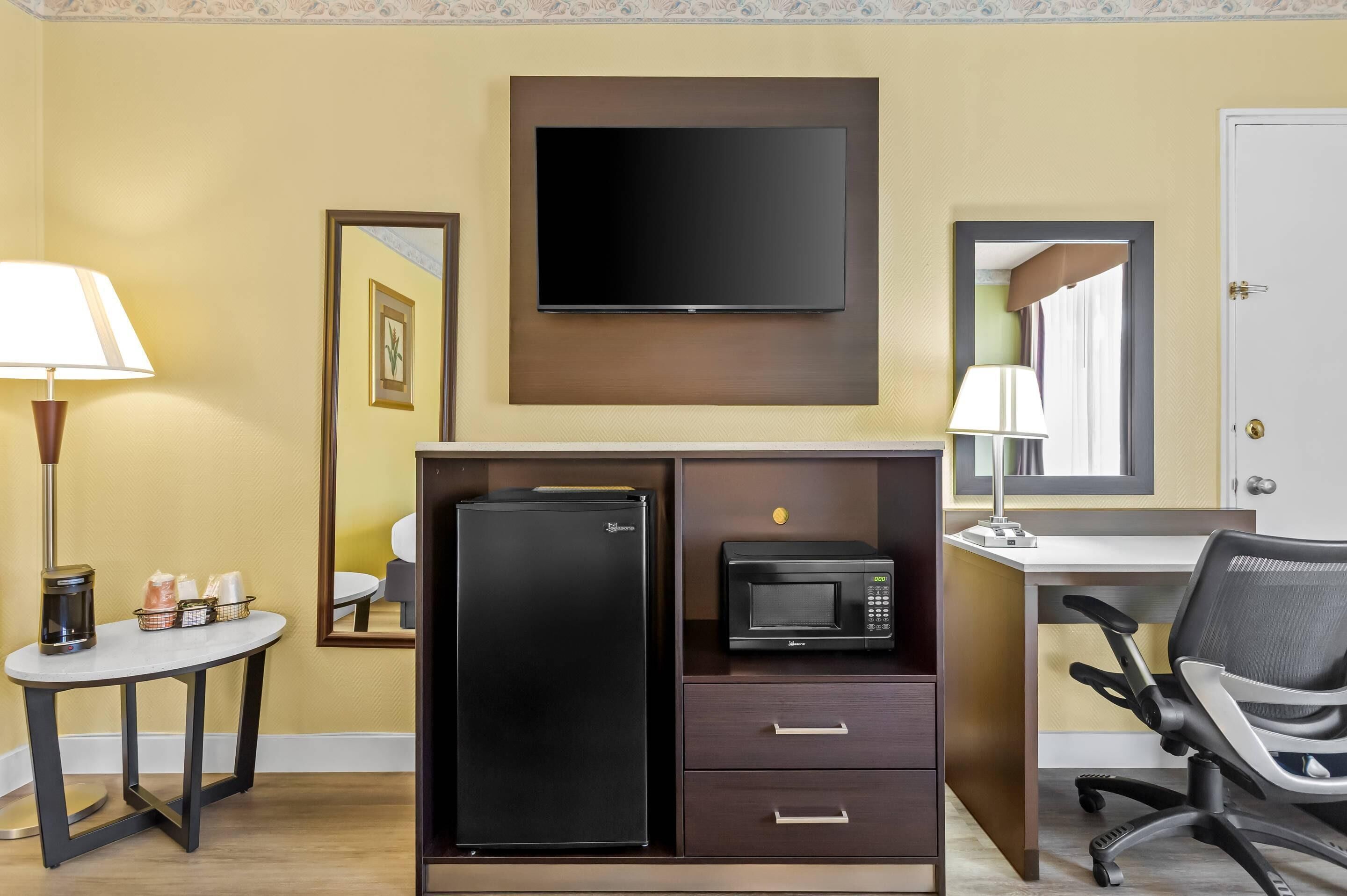 Une chambre d’hôtel équipée d’un téléviseur à écran plat fixé au mur, d’un réfrigérateur, d’un four à micro-ondes, d’un bureau avec une chaise et d’une petite table avec une lampe et quelques objets.