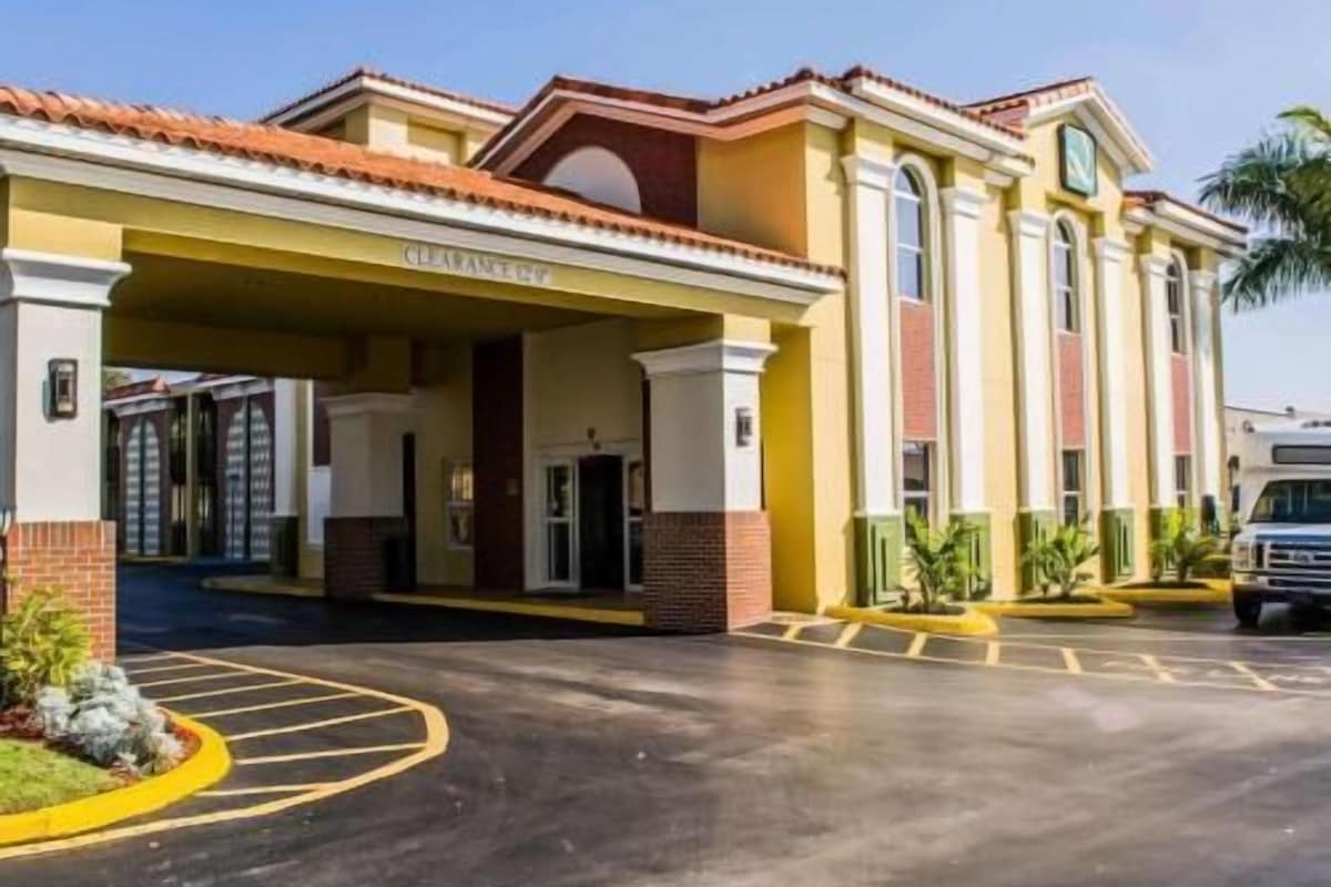 Entrada de hotel con área cubierta, estacionamiento y un vehículo al fondo.