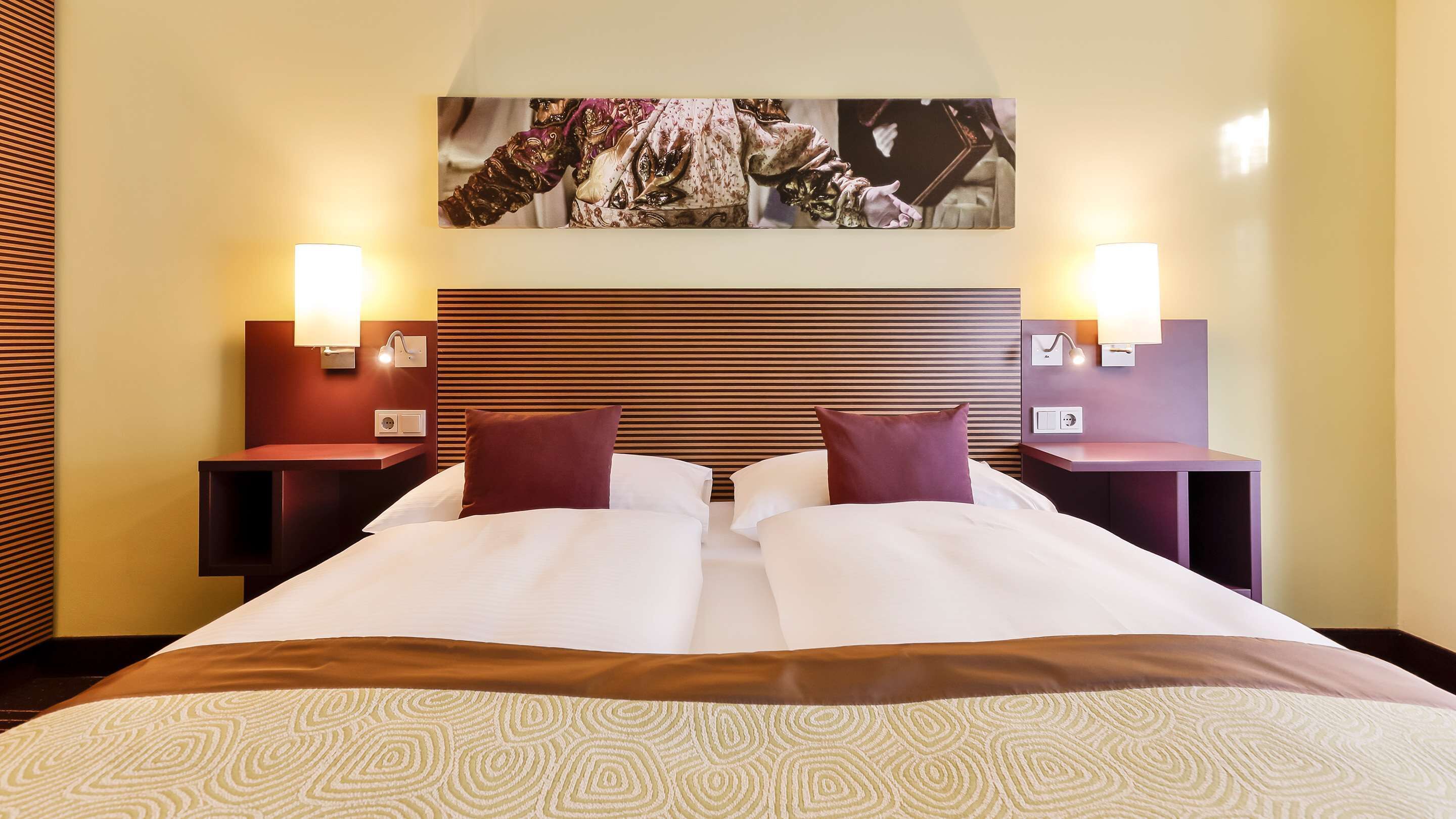 Una habitación de hotel con una cama grande, dos mesitas de noche, un cabecero con líneas horizontales y lámparas fijadas a la pared.
