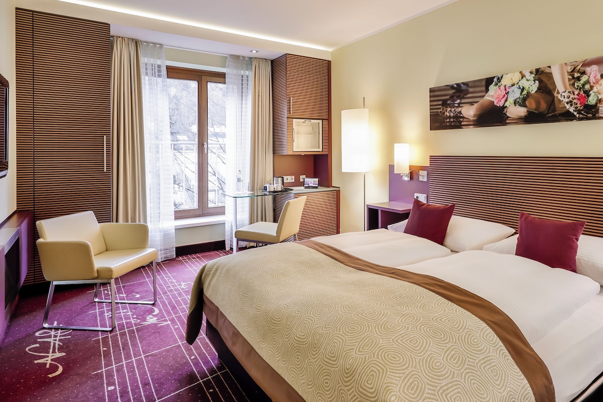 A hotel room with a large bed, a desk, a chair, a TV, and a window with curtains.