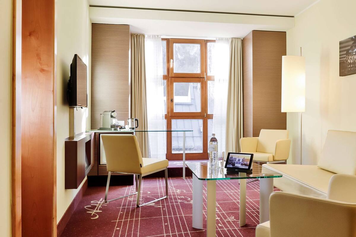 A hotel room with a glass table, two chairs, a sofa, a TV, a window with blinds, and a painting on the wall.