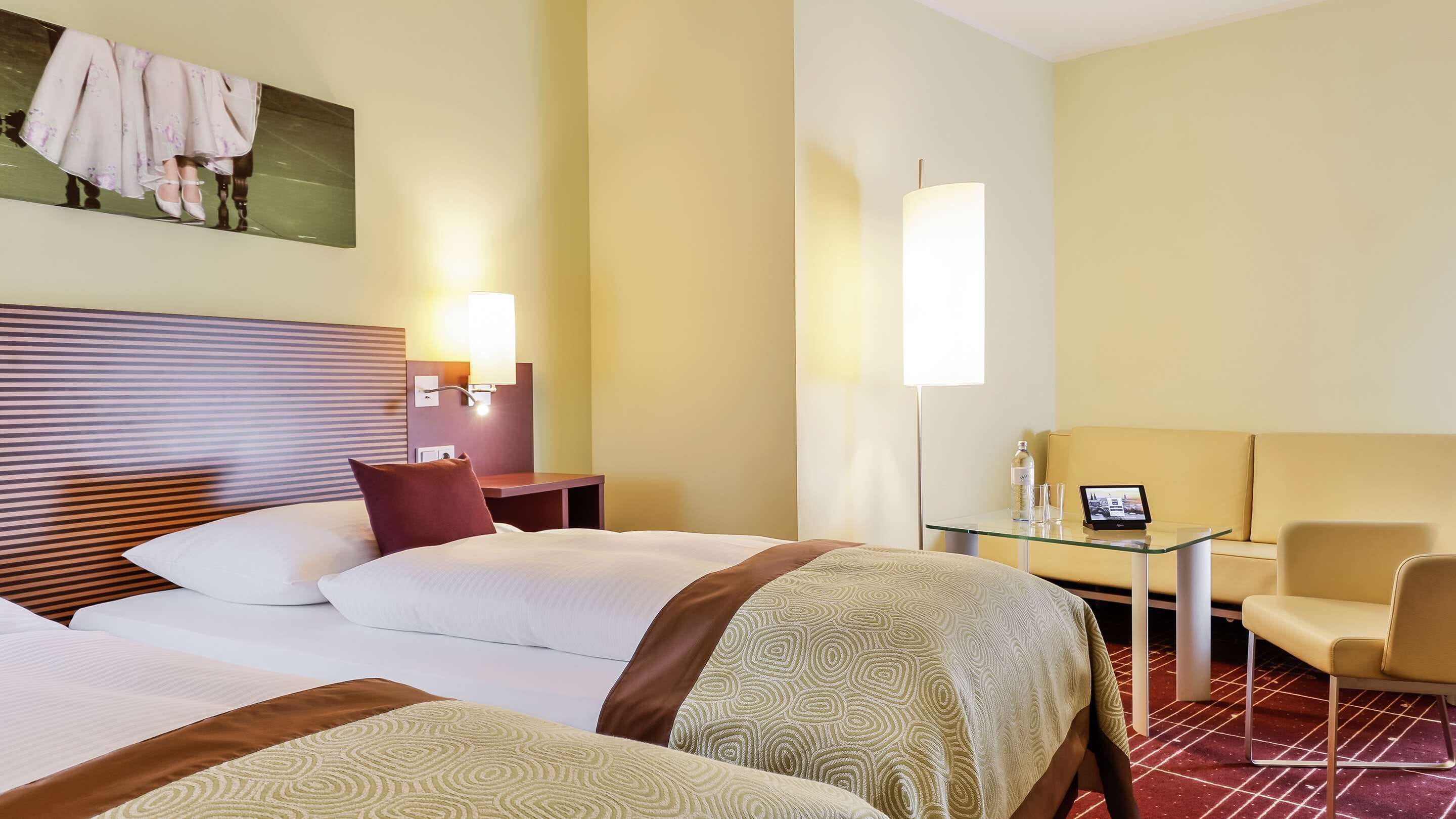 A hotel room with two beds, a desk with a laptop, a sofa, and a wall-mounted TV.