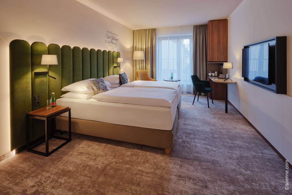 A hotel room with a large bed, a desk, a chair, a TV, and a wall-mounted lamp.