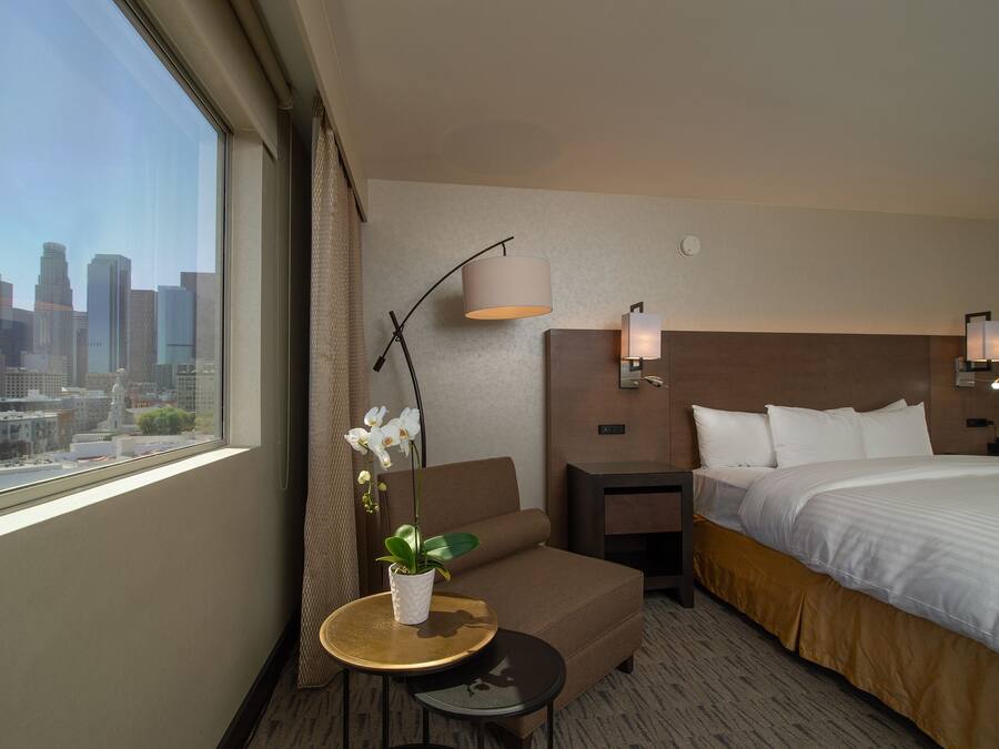 Quarto de hotel com cama, um pequeno sofá, mesa de cabeceira, luminária, orquídea em vaso e vista para o horizonte da cidade.