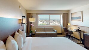 Une chambre d’hôtel avec un grand lit, un canapé, un bureau et une vue sur le paysage urbain, visible à travers une grande fenêtre.
