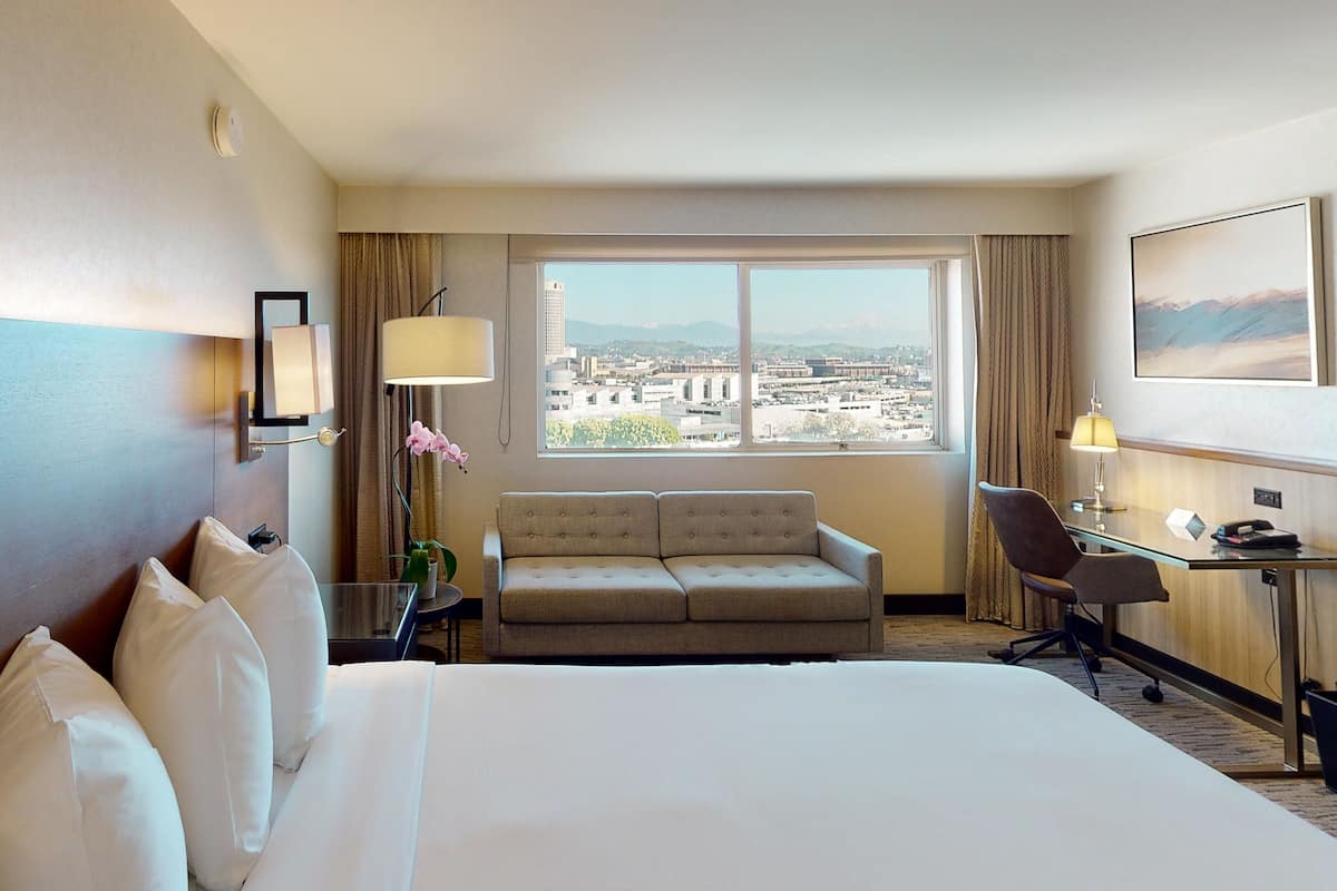 A hotel room with a large bed, a sofa, a desk, and a view of a cityscape through a large window.
