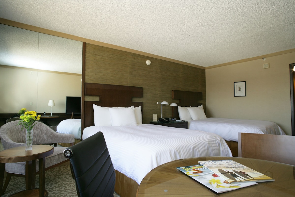 A hotel room with two beds, a desk, a chair, a small table with a vase of flowers, and a magazine on the table.