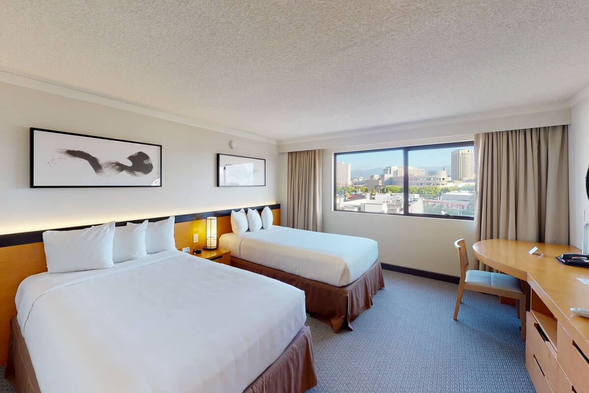 A hotel room with two beds, a desk, a chair, and a large window offering a city view.
