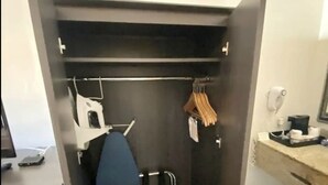 Una habitación pequeña con una puerta oscura, un escritorio con computadora y cargador, un perchero con ropa y una plancha.
