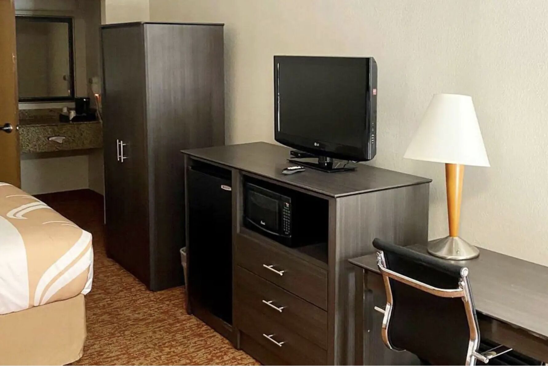 Una habitación de hotel compacta con una cama, un escritorio con televisor, un refrigerador pequeño y una lámpara.