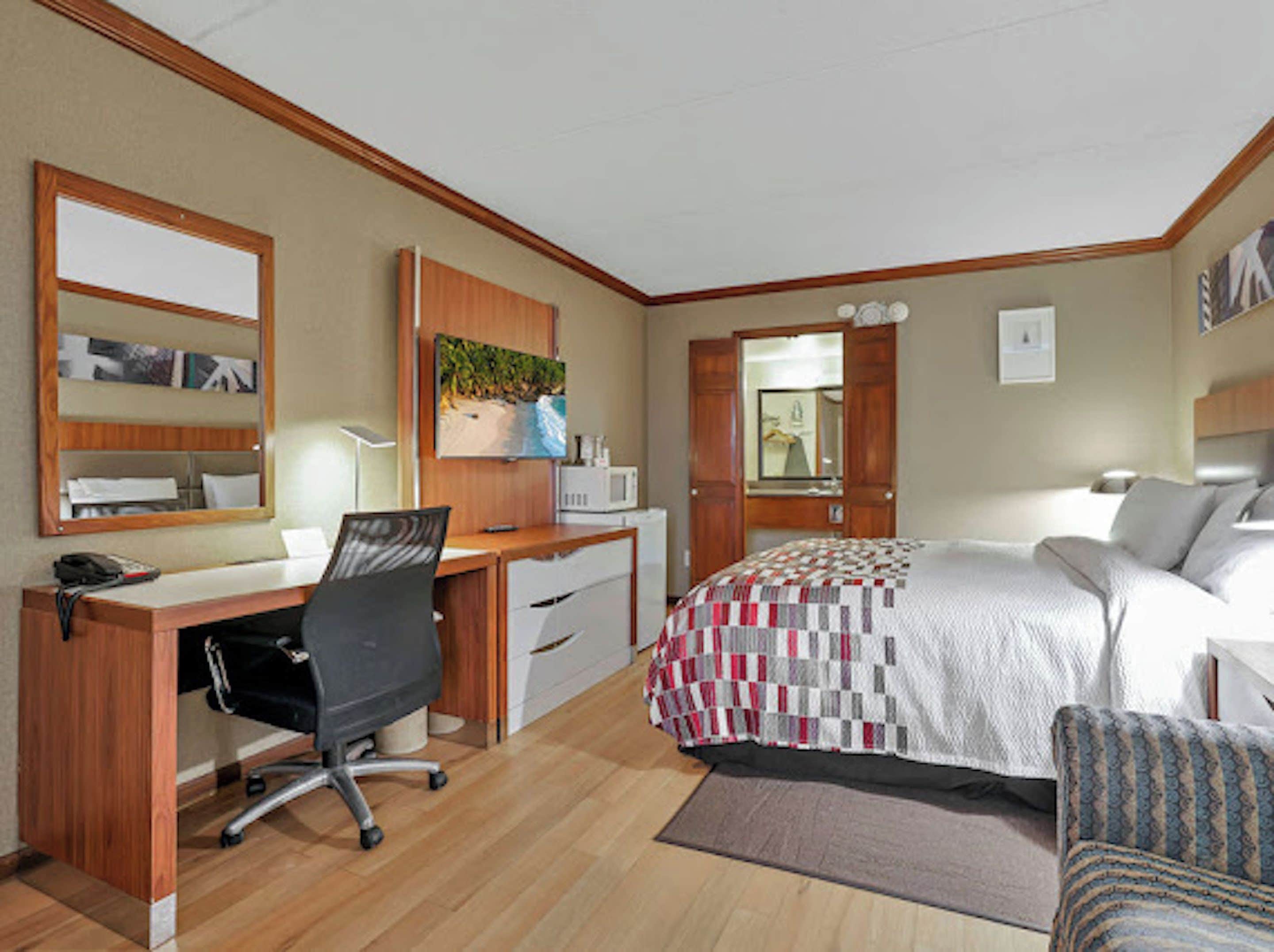 Een hotelkamer met een houten bureau, een stoel, een bed met een sprei, een spiegel en een deur naar een badkamer.