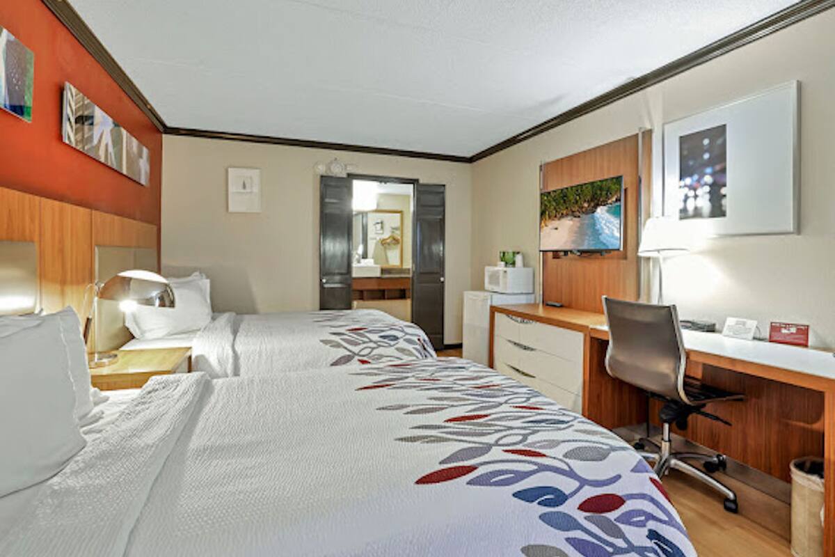 A hotel room with two beds, a desk, a chair, a microwave, and a television.